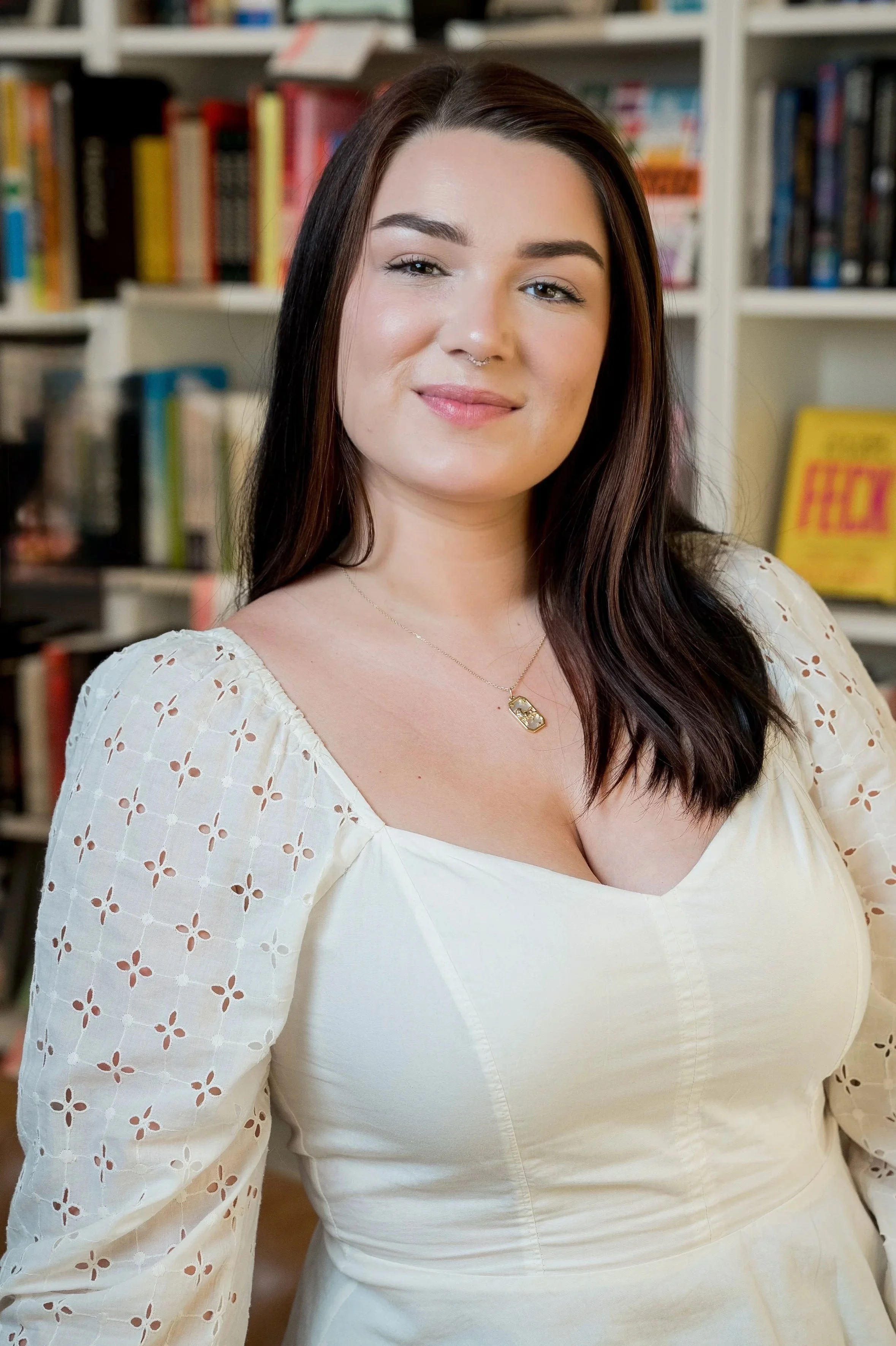 Close-up of a young woman with long dark hair, wearing a white dress with eyelet pattern on the sleeves, standing in front of bookshelves.