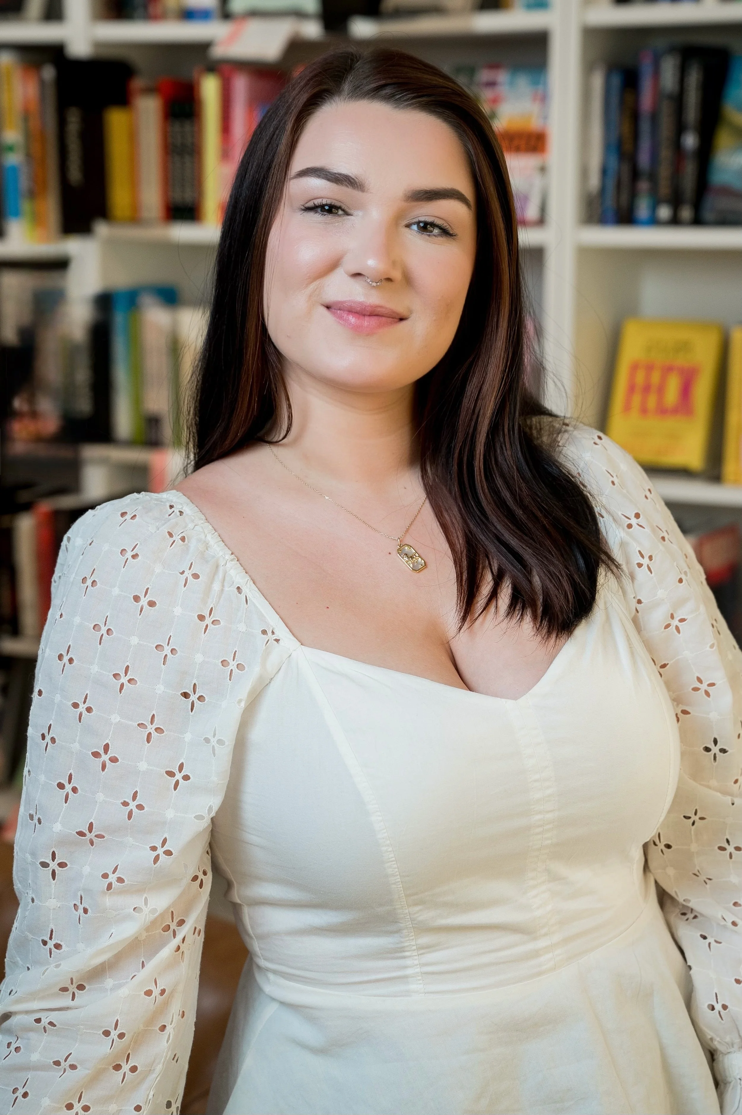 A woman with long dark hair, wearing a white dress with eyelet sleeves and a delicate necklace, standing in front of a bookshelf filled with colorful books.