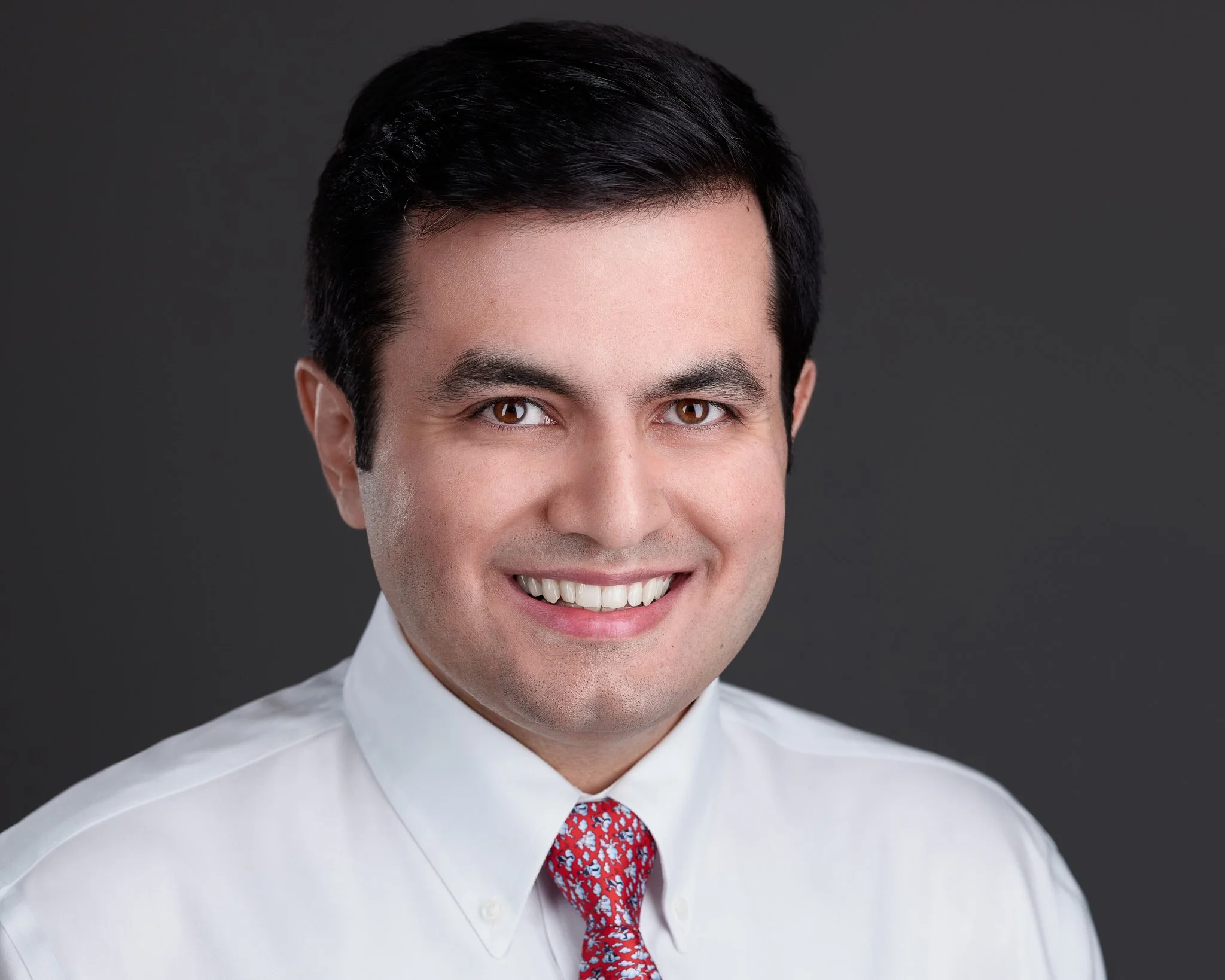 A man with dark hair, brown eyes, and a light skin tone is smiling and wearing a white shirt with a red tie that has a white floral pattern, set against a dark gray background.