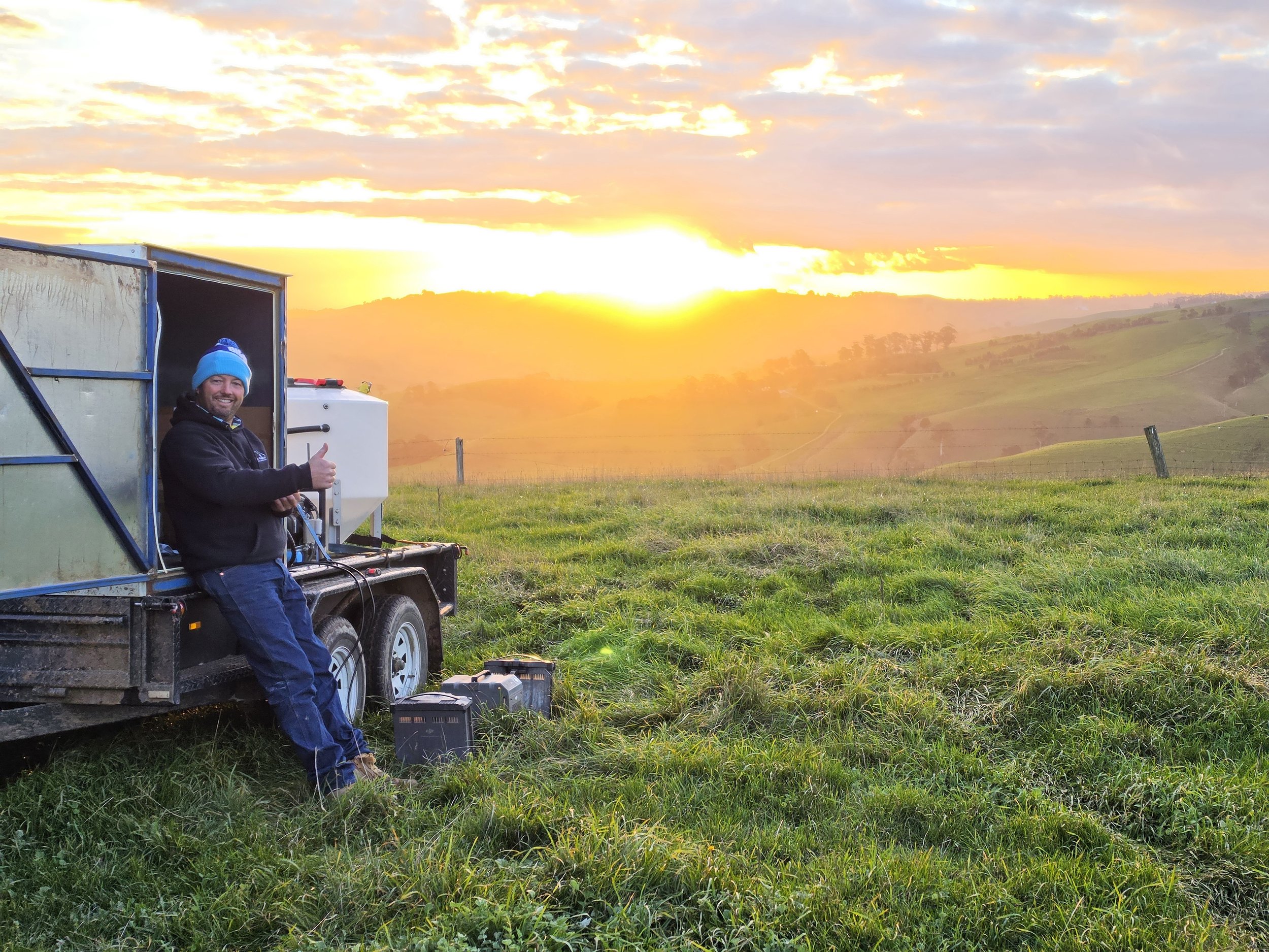 A man giving a thumbs-up while standing next to a trailer with a sunrise in the background over rolling hills and grassy fields.