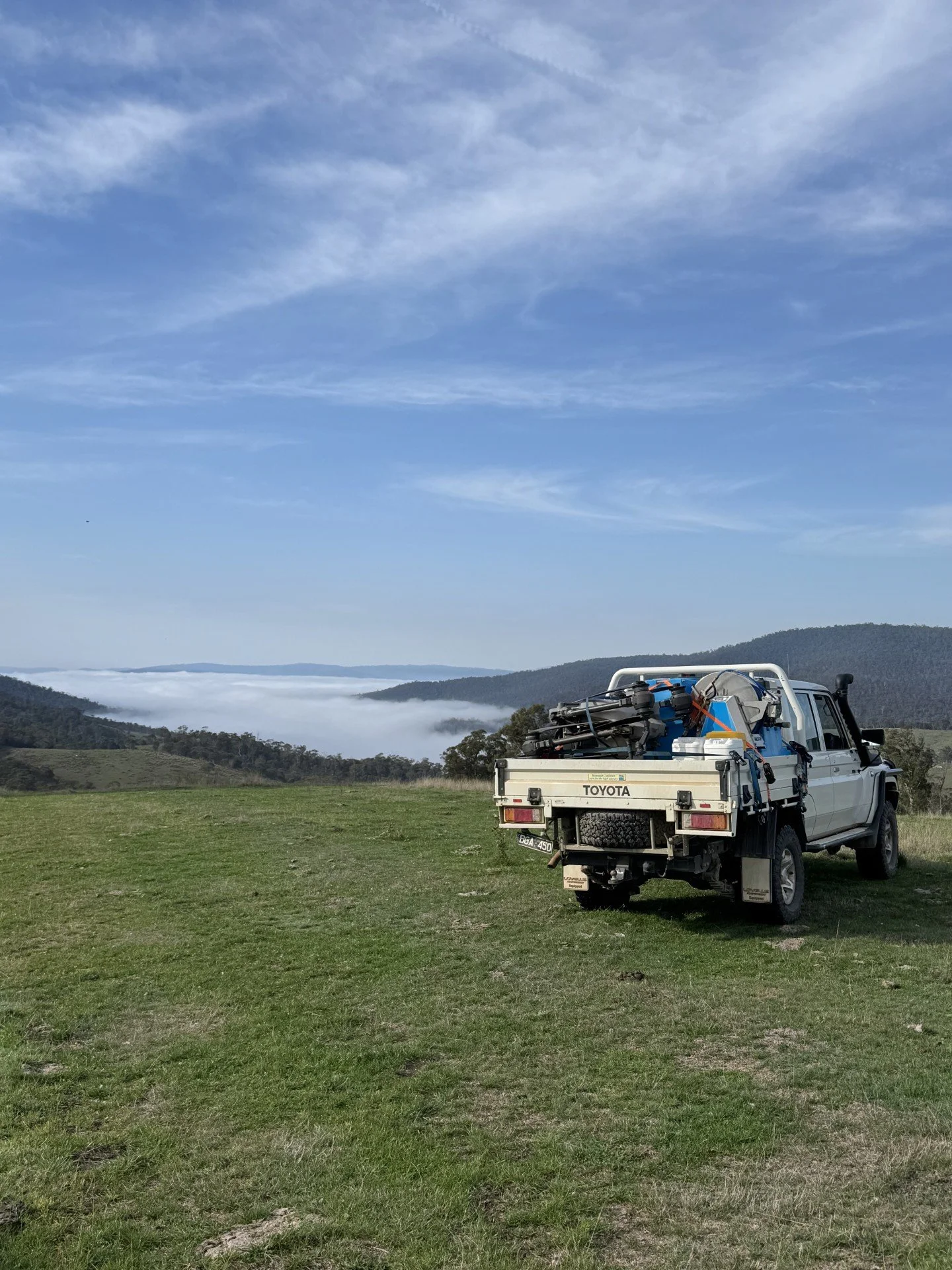 A white Toyota pickup truck parked on a grassy field with open land, hills, and mountains in the background under a partly cloudy sky.