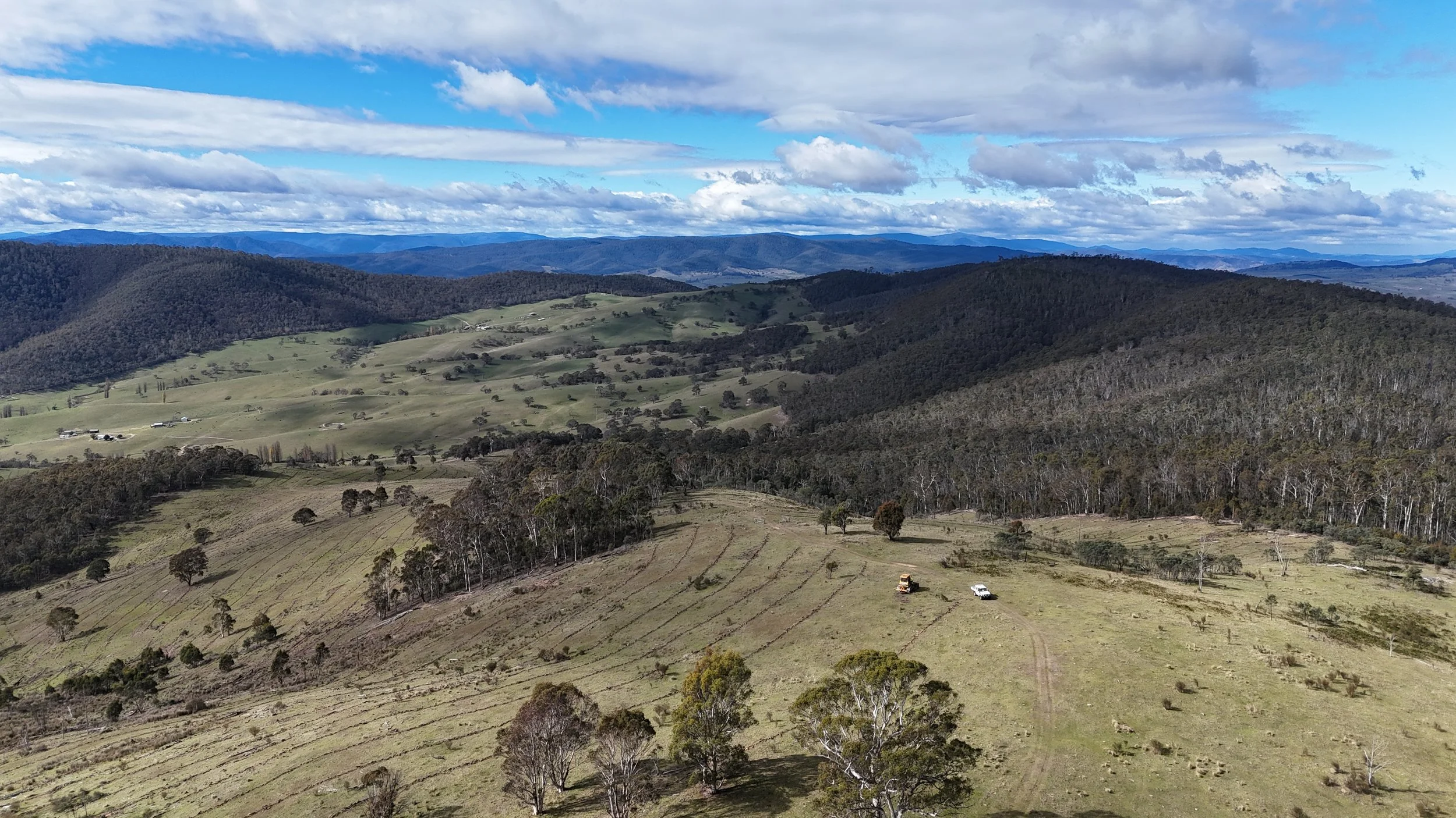A scenic landscape with rolling hills, patches of green grass, scattered trees, and a partly cloudy sky with blue patches, with some small structures and vehicles visible in the distance.