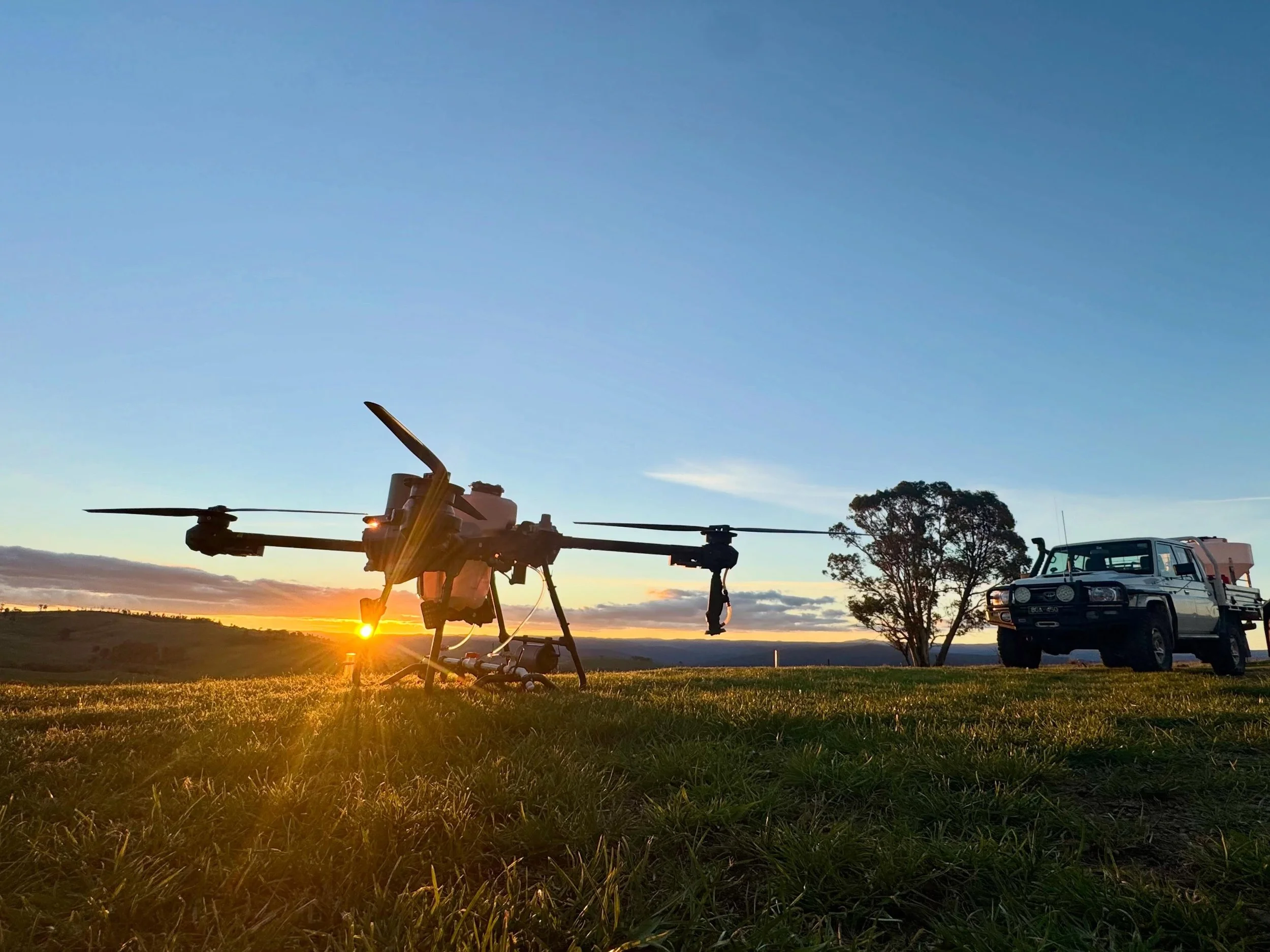 A drone on the grass with a sunset in the background, along with a police vehicle and trees.