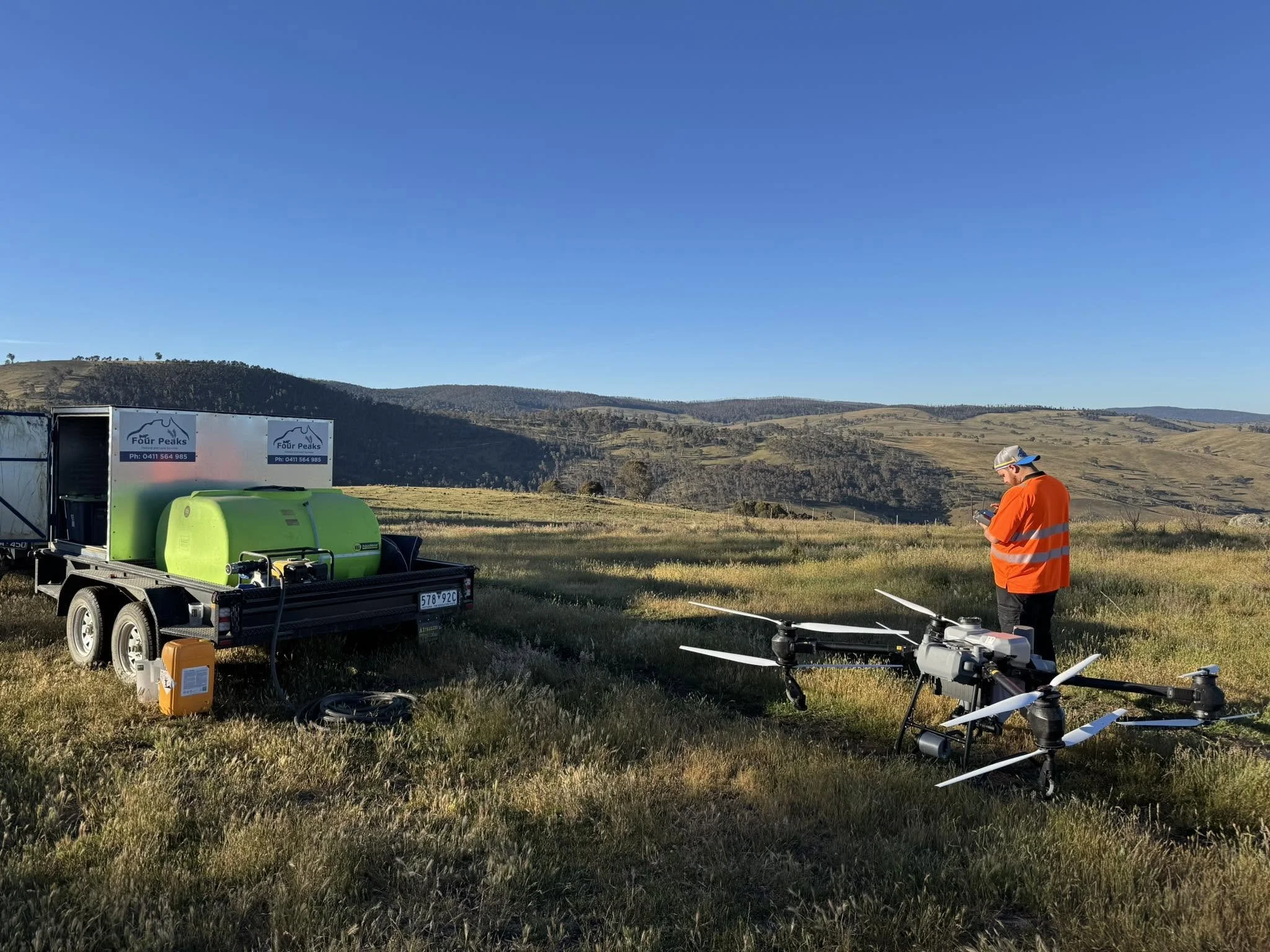 A person wearing an orange safety vest and a cap is standing outdoors near a drone, with a trailer carrying equipment like a spray tank labeled 'Four Peaks'. The background features rolling hills under a clear blue sky.