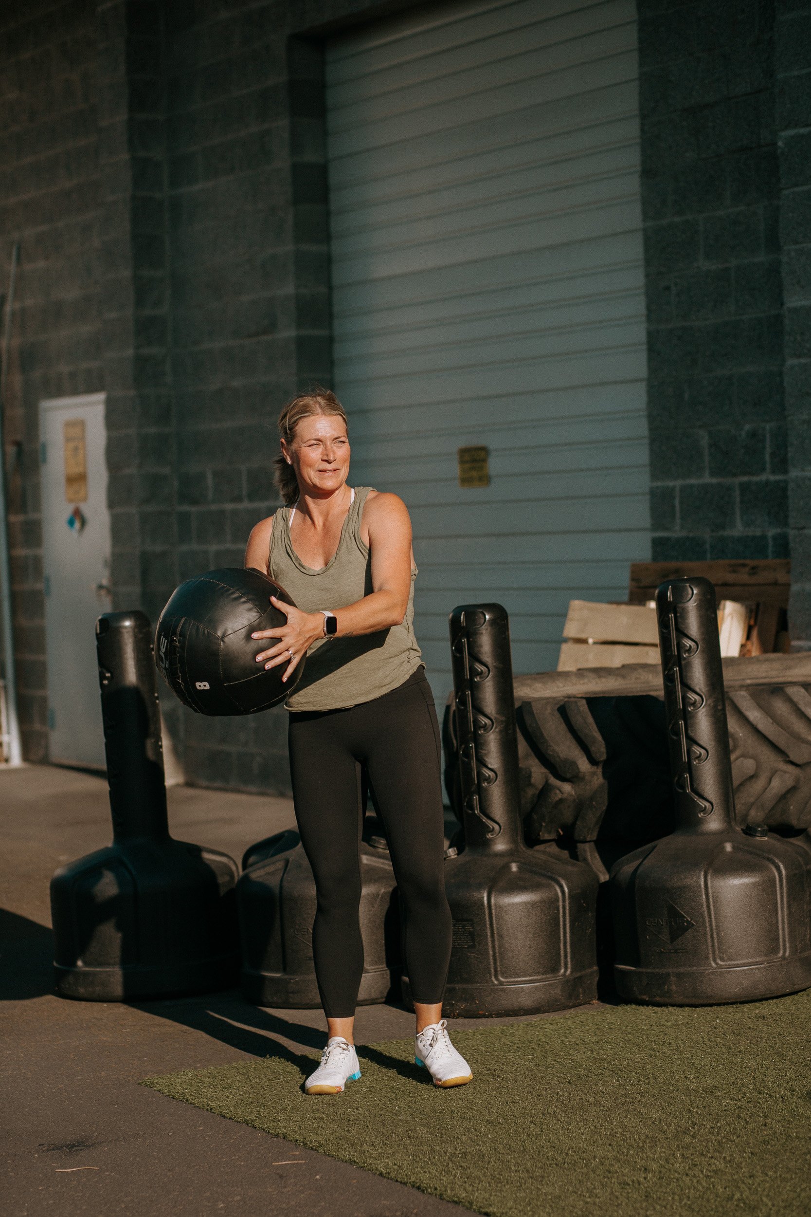 Dropgym member exercising outside of the gym with a medicine ball in her hands at a gym or training facility with large black tires and a gray wall in the background.