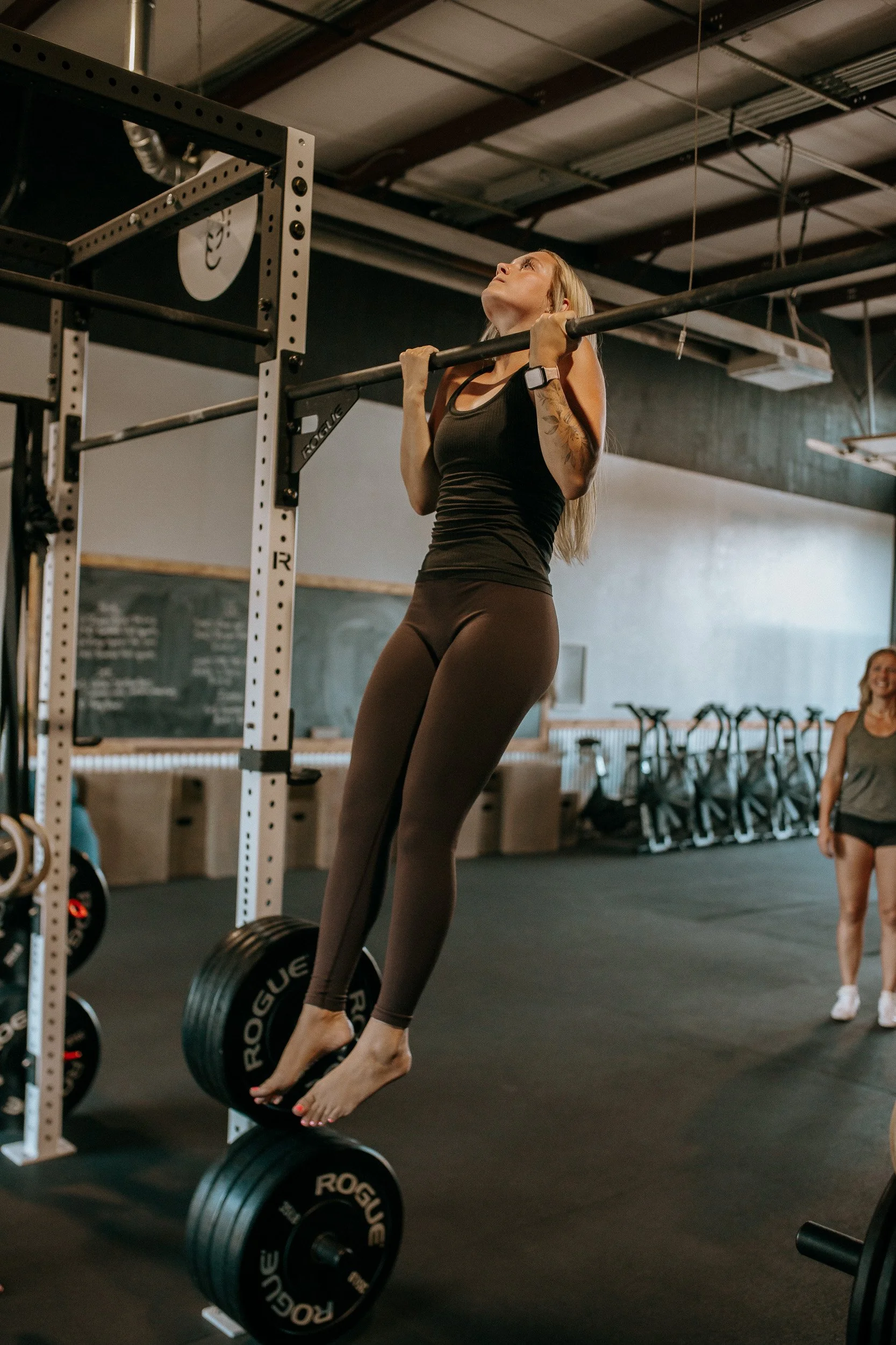 Dropgym member doing a pullup, wearing brown leggings and a black tank top. Other members watch and cheer her on.