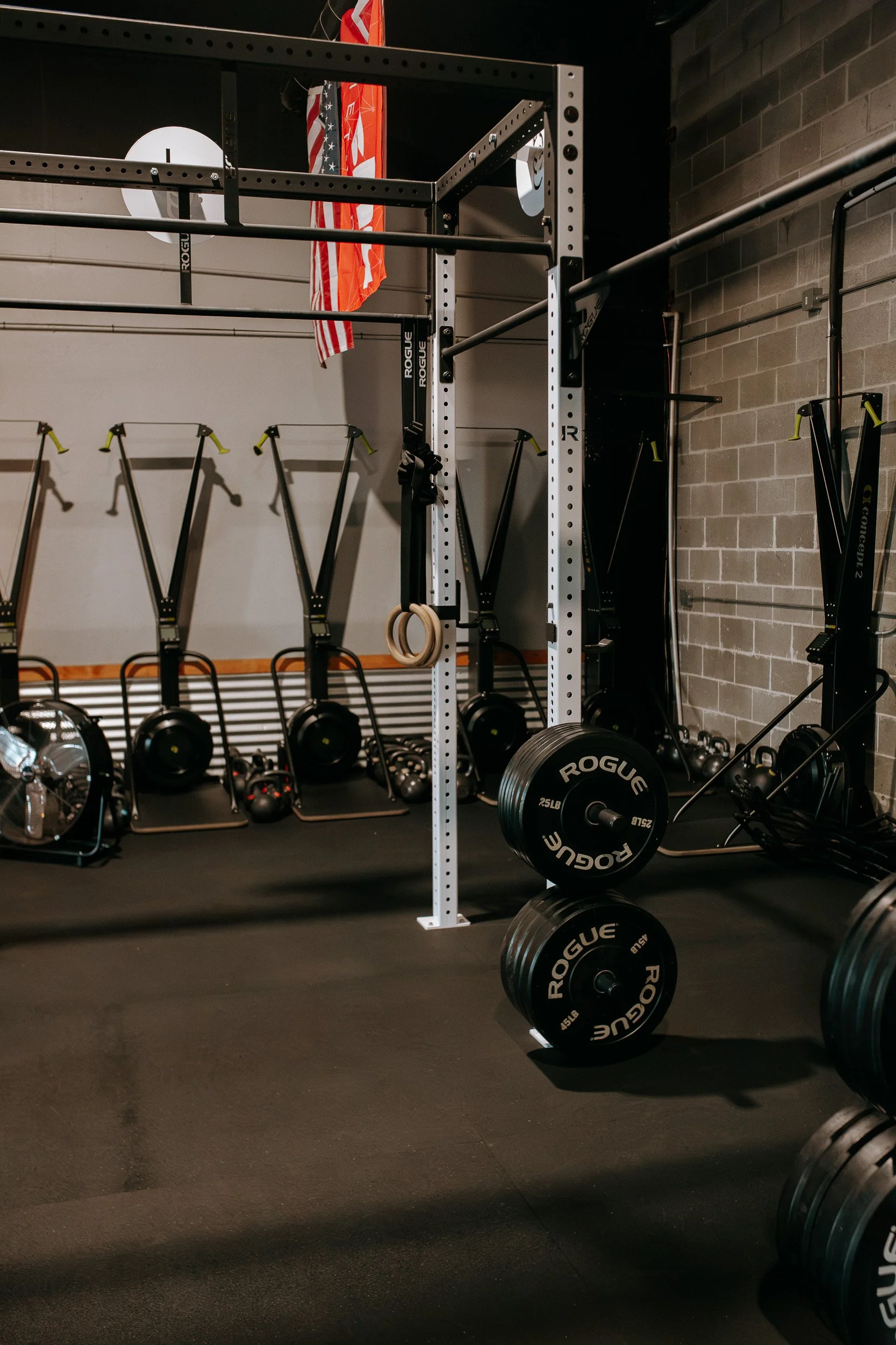 Dropgym's interior with functional fitness equipment including barbells, weight plates, gymnastic rings, and resistance bands, set against a black rubberized floor and brick wall, with flags hanging in the background.