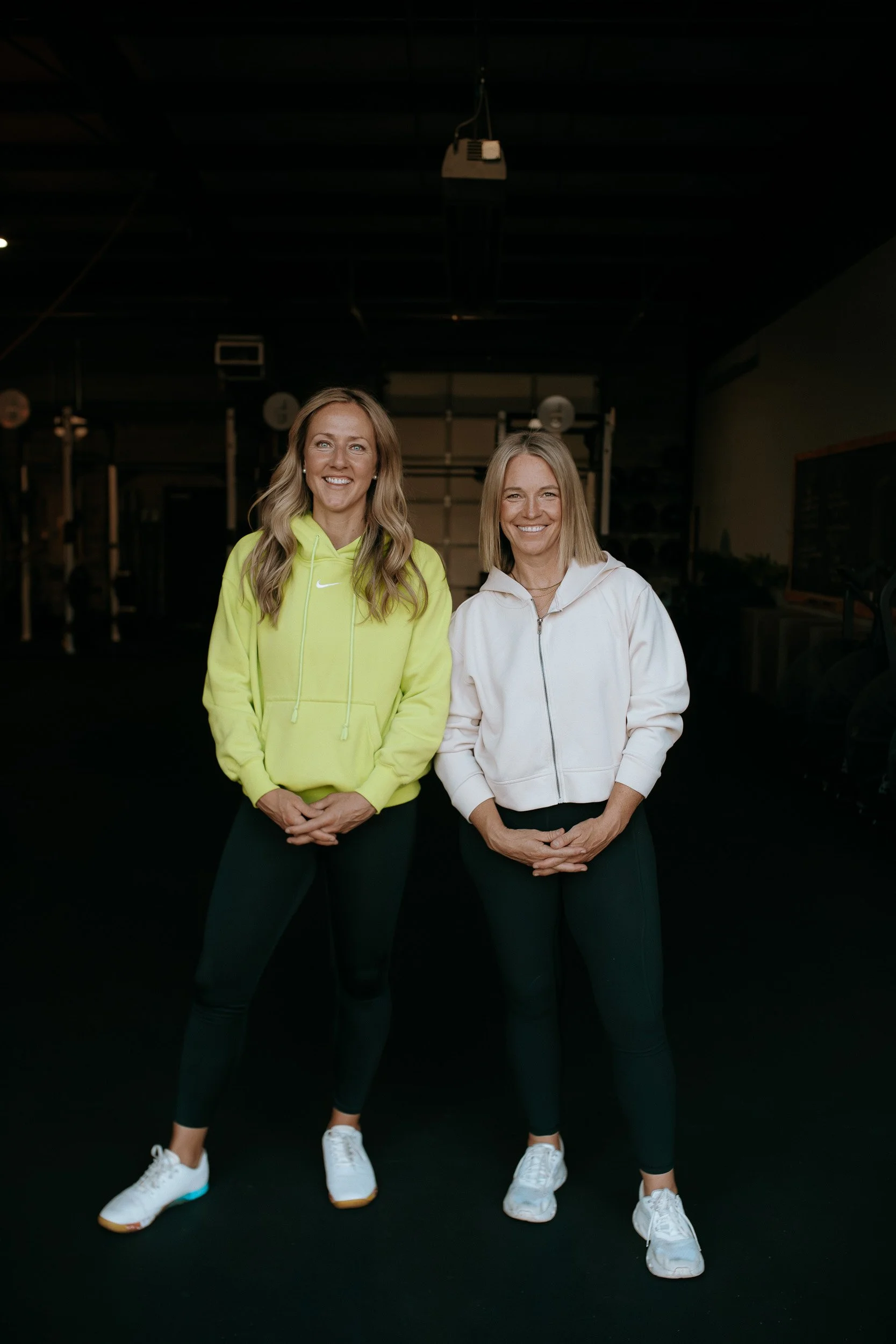 Lu and Shantell, co-owners of Dropgym Medford, smiling at the camera. Both are wearing athletic clothing and sneakers, with a dark background featuring gym equipment.