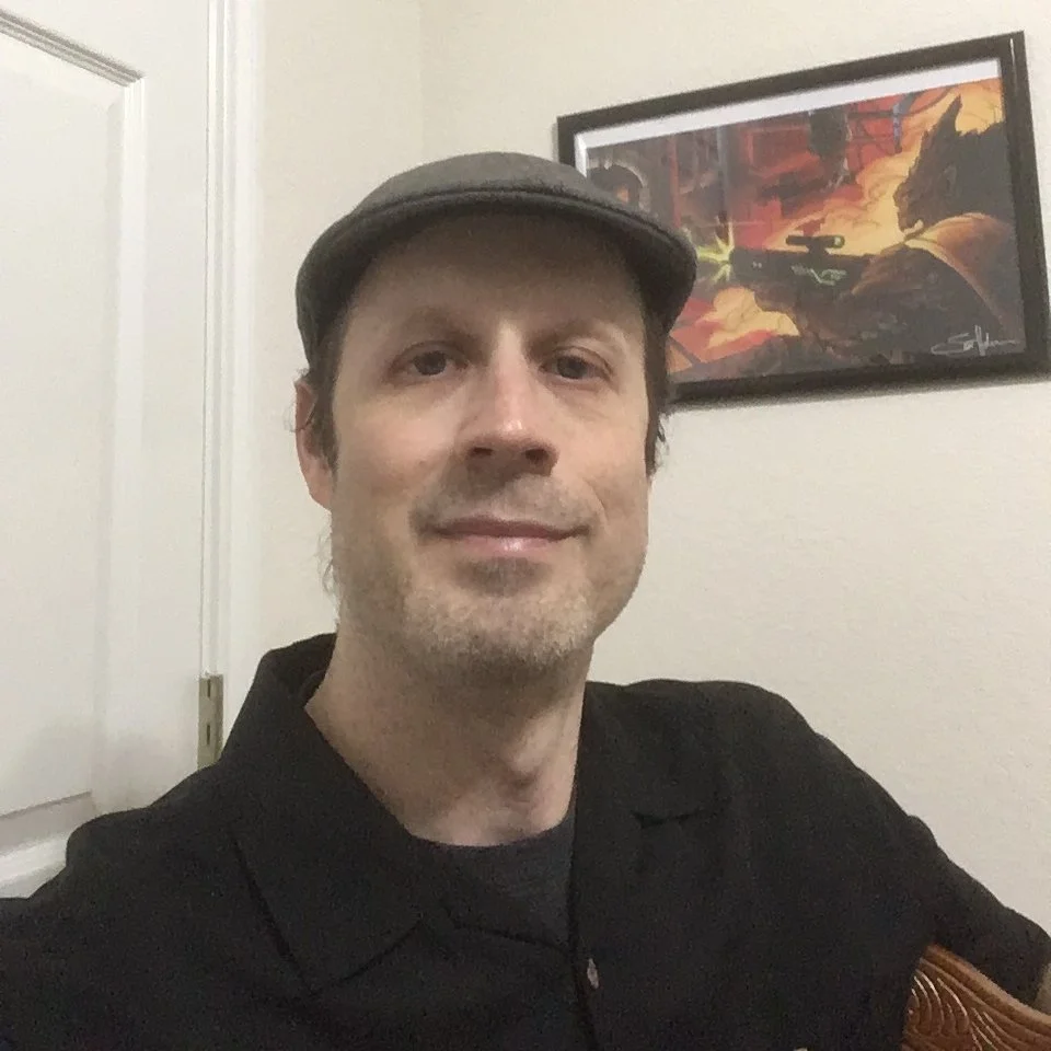A man wearing a gray flat cap and black shirt, smiling slightly, sitting in a room with a framed comic book-style poster from a sci-fi audio drama called Caalo Xan.