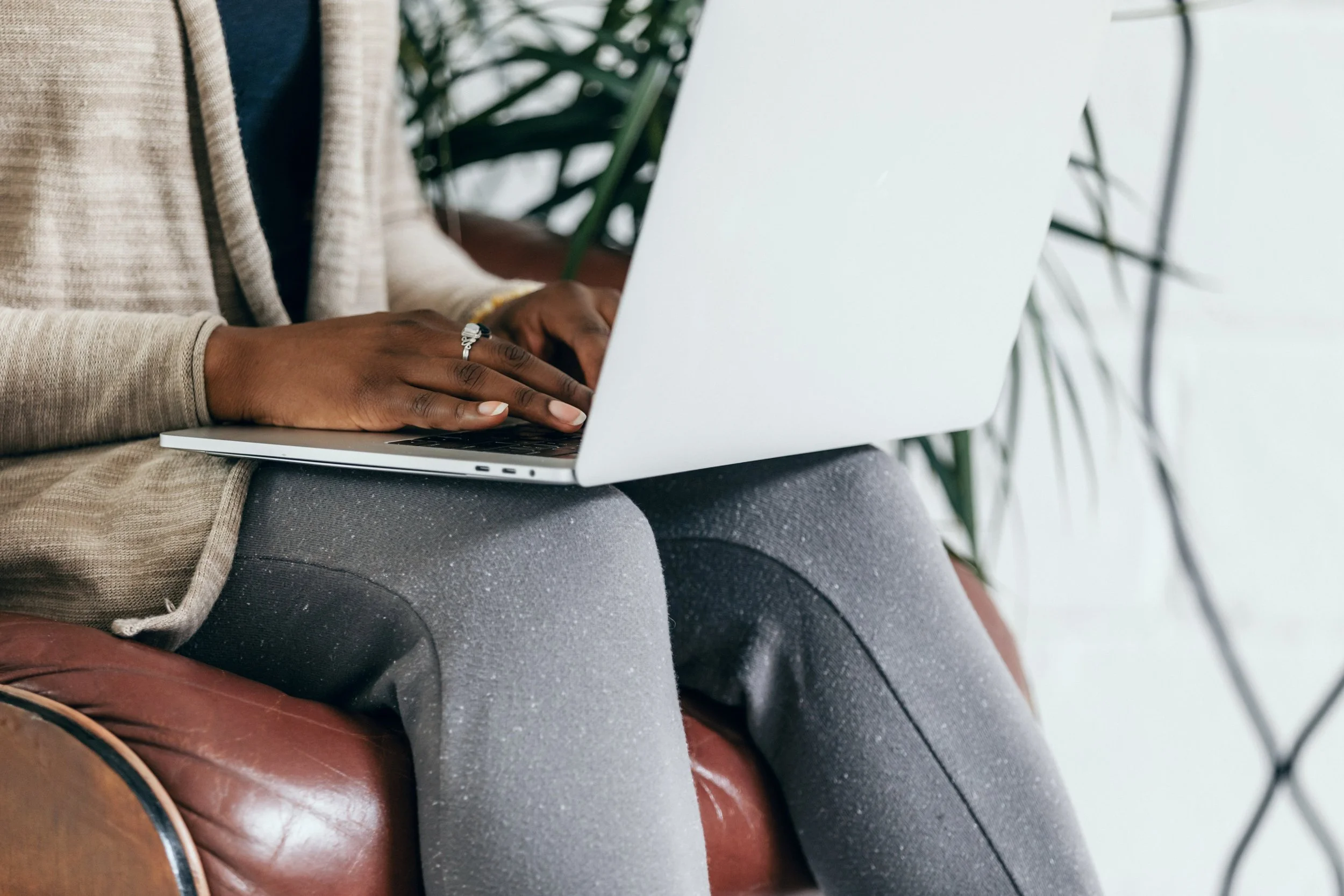 A person sitting on a brown leather chair working on a silver laptop. The individual is wearing a beige cardigan and gray pants, with a silver ring on their finger. There are green plants in the background.