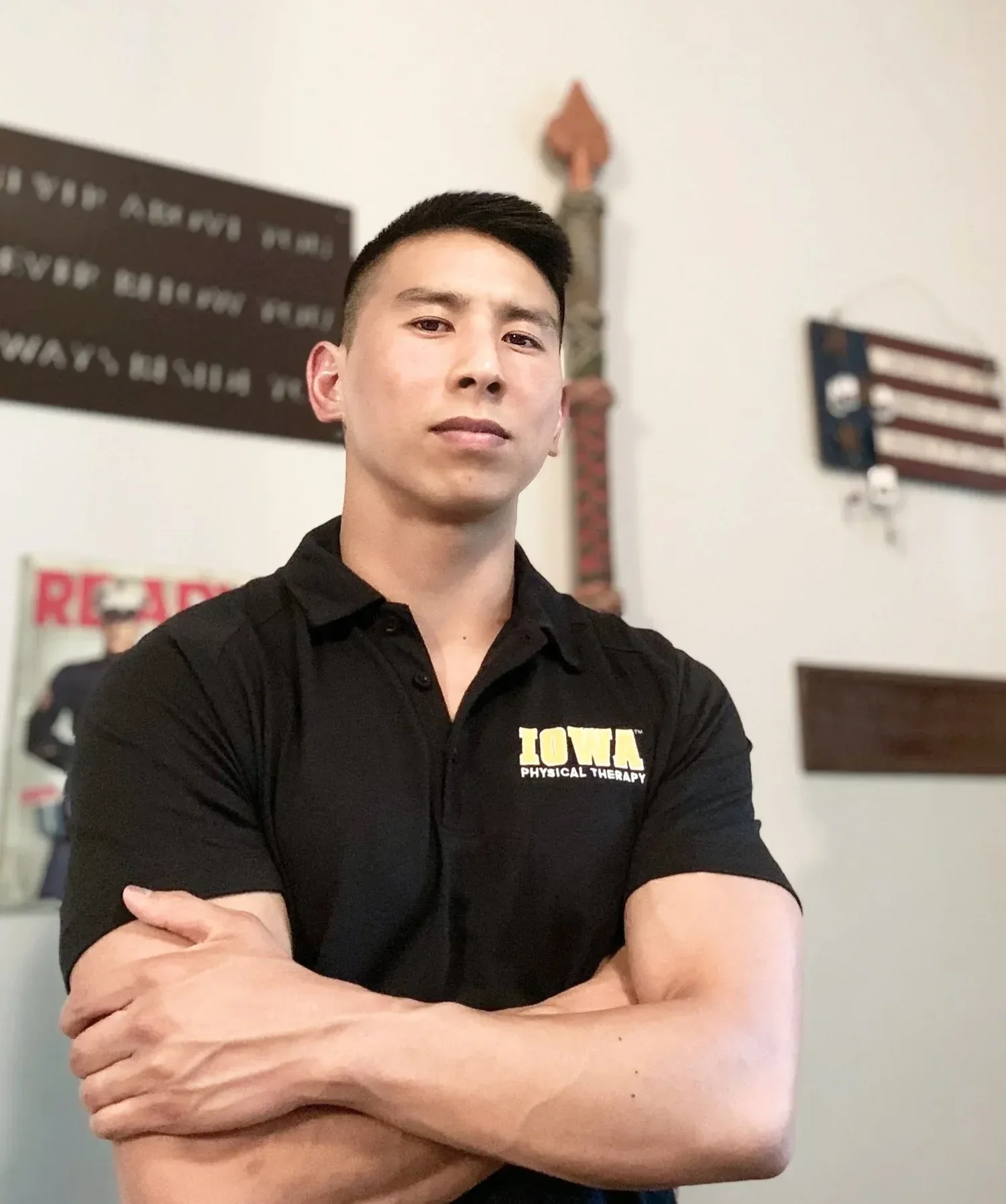 Young man with short black hair and light skin, wearing a black polo shirt with yellow text that reads 'IOWA Physical Therapy,' standing indoors with arms crossed, in front of wall decorations including a sign, a wooden American flag, and a tall carved wooden decoration.