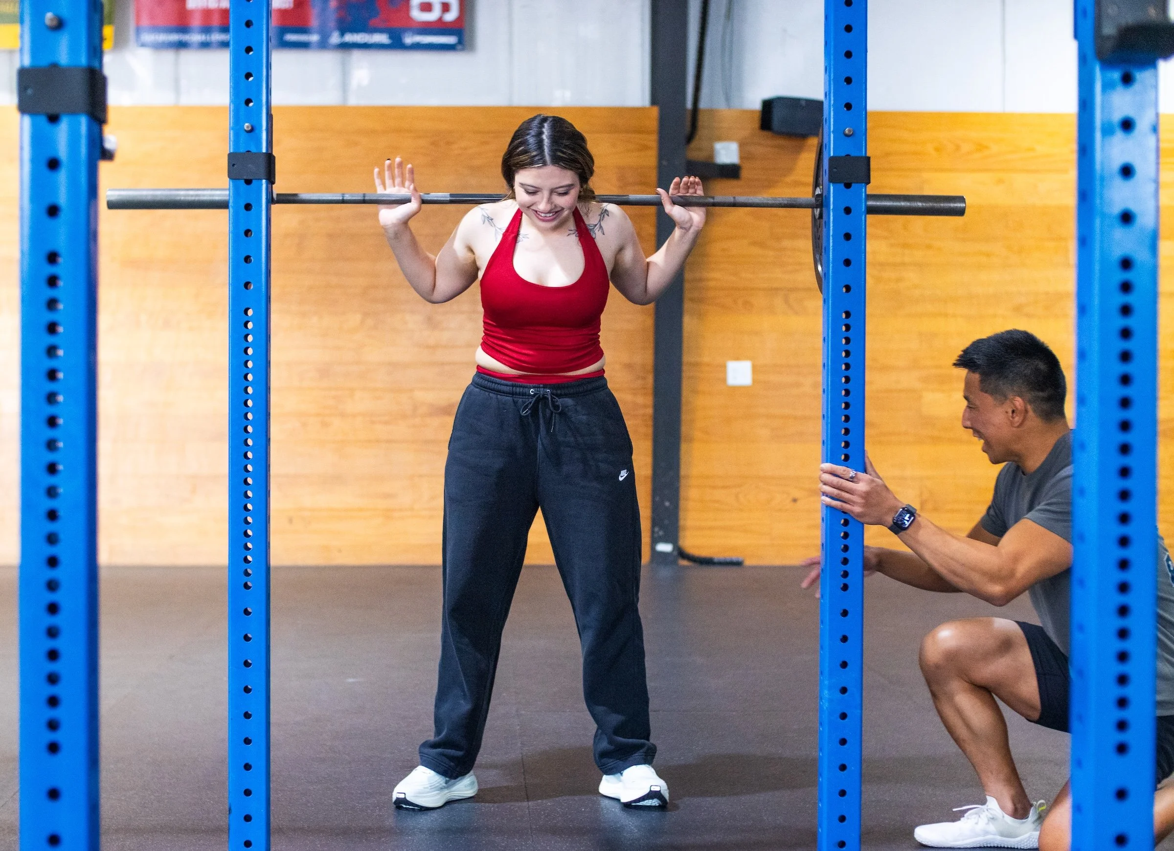 A woman in a red sports top and black sweatpants is in a gym, smiling as she prepares to lift a barbell. A man crouches nearby, helping her with the barbell rack. Blue gym equipment frames the scene.