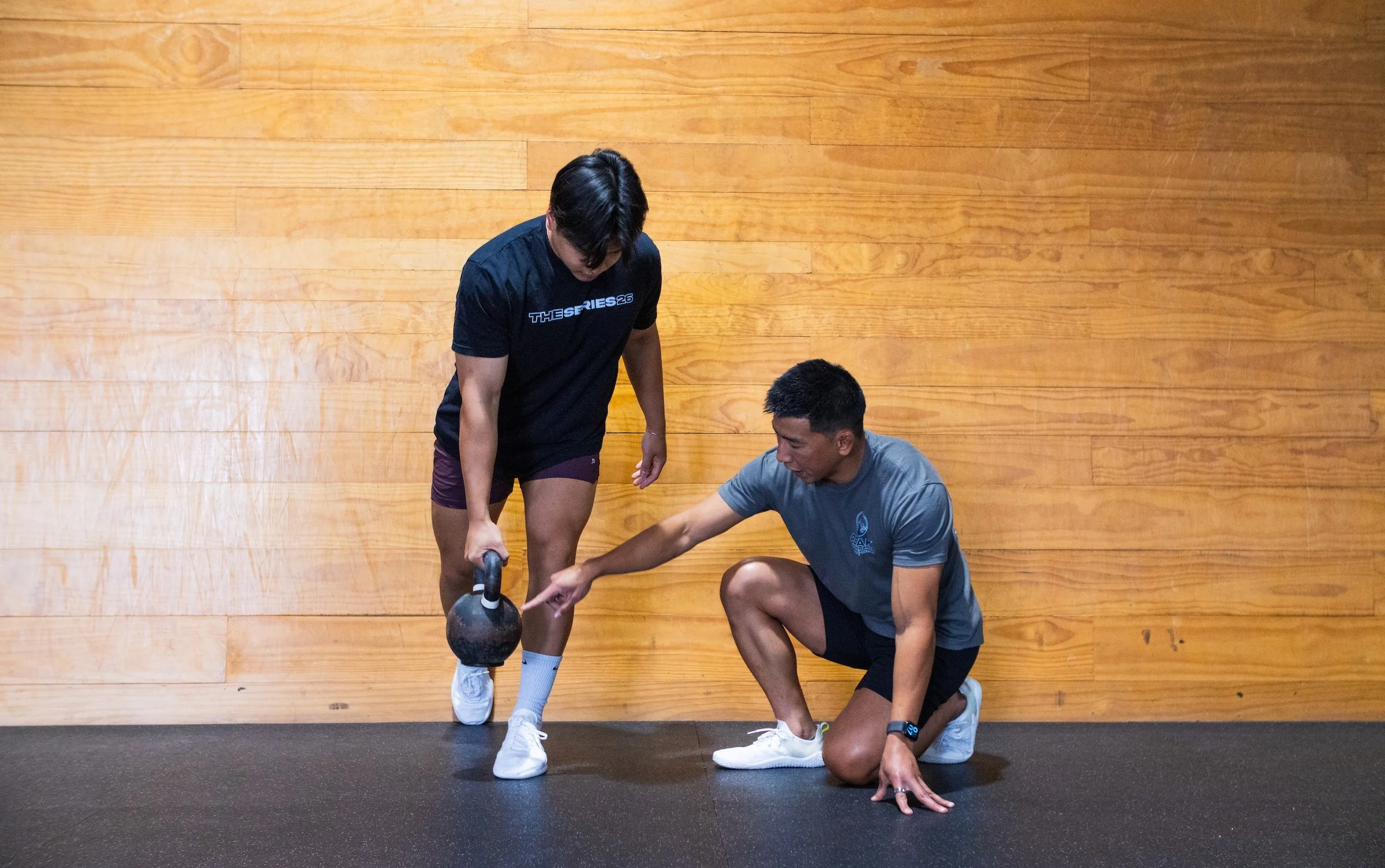 Two men in athletic clothing are performing a workout with a kettlebell in a gym with a wooden wall. One man is standing, holding the kettlebell with one hand while the other man is kneeling and pointing at the kettlebell.