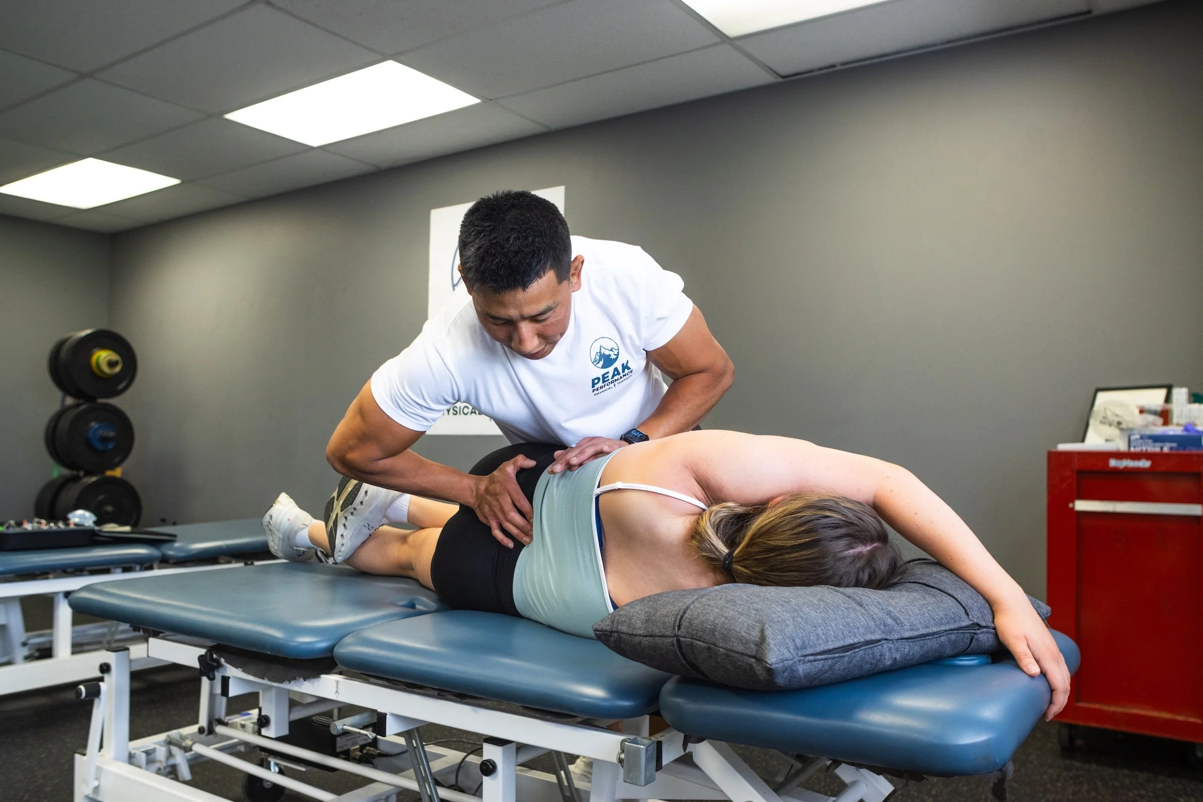 A chiropractor performing an adjustment on a woman lying face down on a treatment table. The setting is a professional clinic with gray walls, a red cabinet, and gym equipment visible in the background.