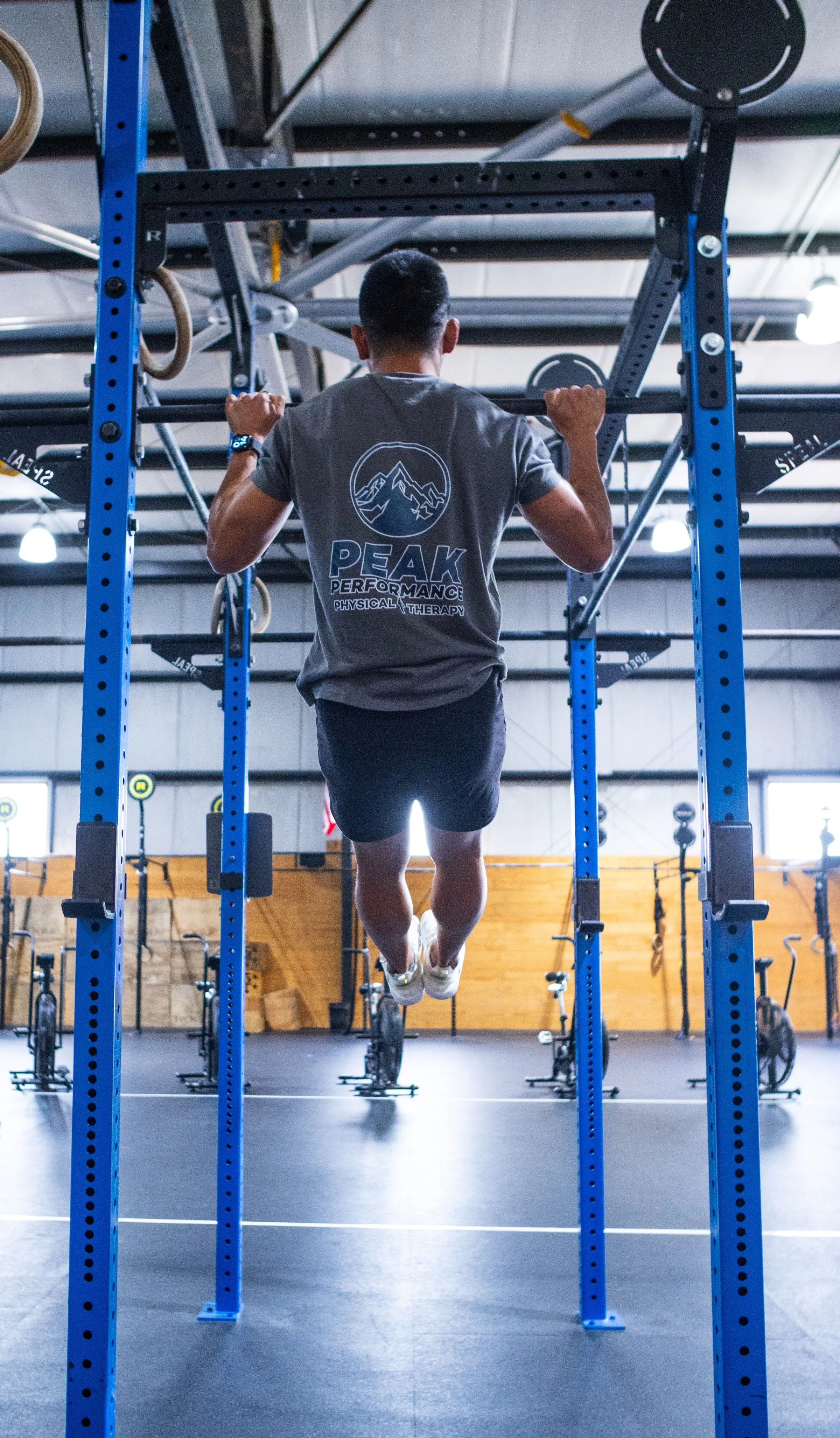 Man performing a pull-up exercise in a gym, wearing a gray T-shirt with the logo 'PEAK PERFORMANCE PHYSICAL THERAPY' on the back, and black shorts.