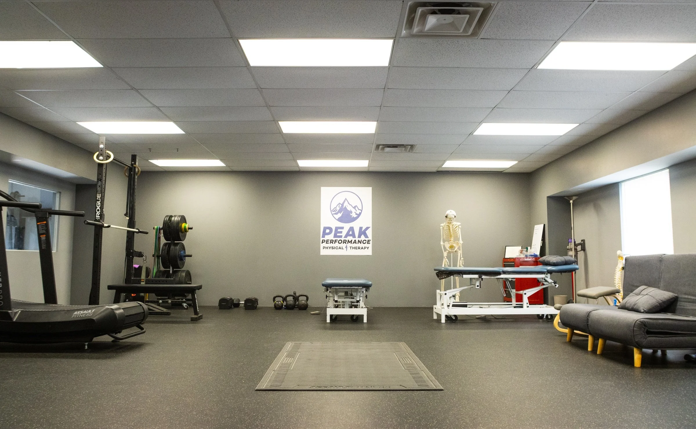 Interior of a physical therapy or fitness center with gym equipment, exercise mats, a skeletal model, and a sign that reads 'Peak Performance Physical Therapy'.