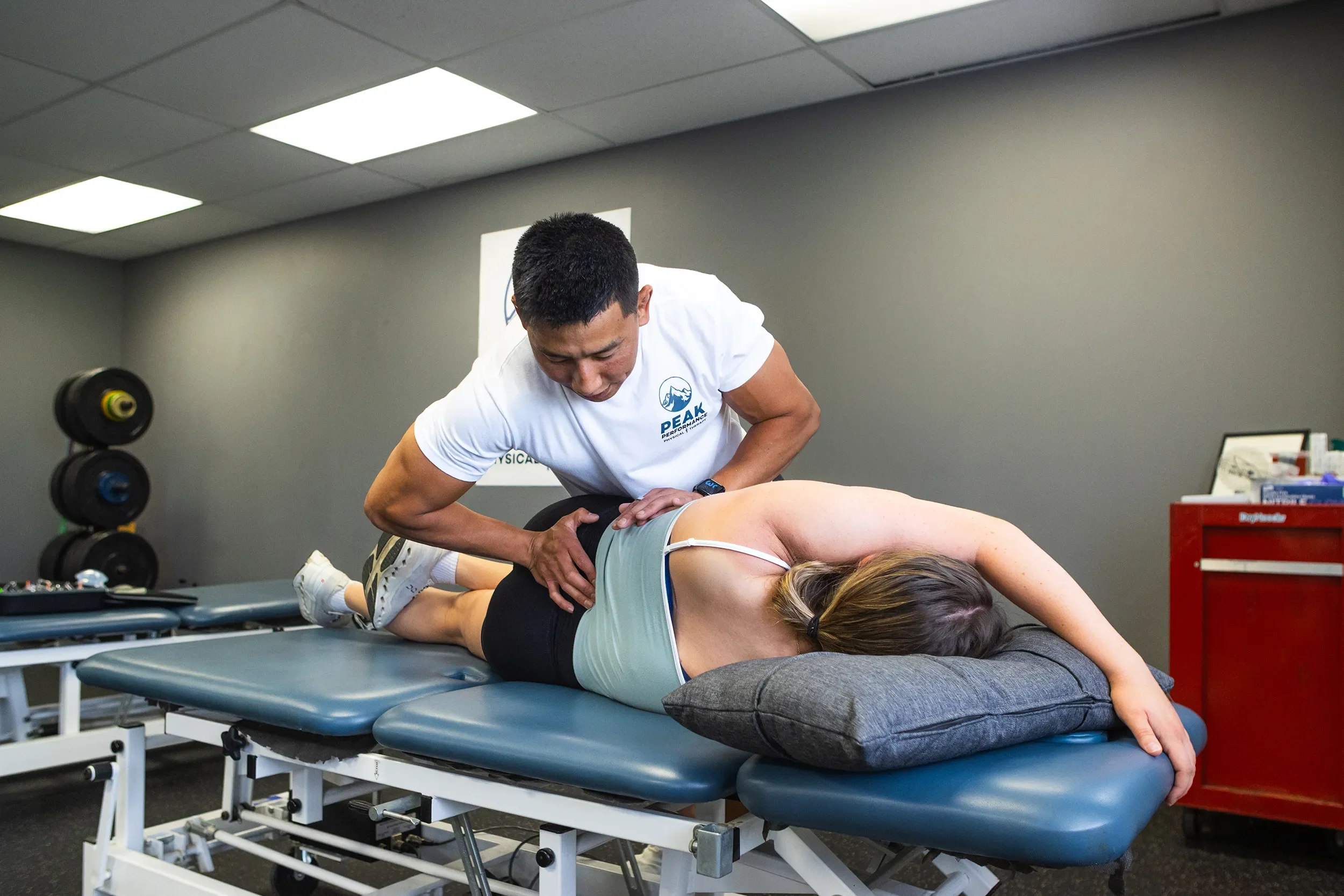 A physical therapist performs a manual therapy technique on a woman lying face down on a treatment table. The woman is wearing a sports bra and shorts. The therapist is wearing a white T-shirt with a logo and text that reads "PEAK Performance". The room has gray walls, a ceiling with fluorescent lights, some weightlifting equipment, and a red medical cart.