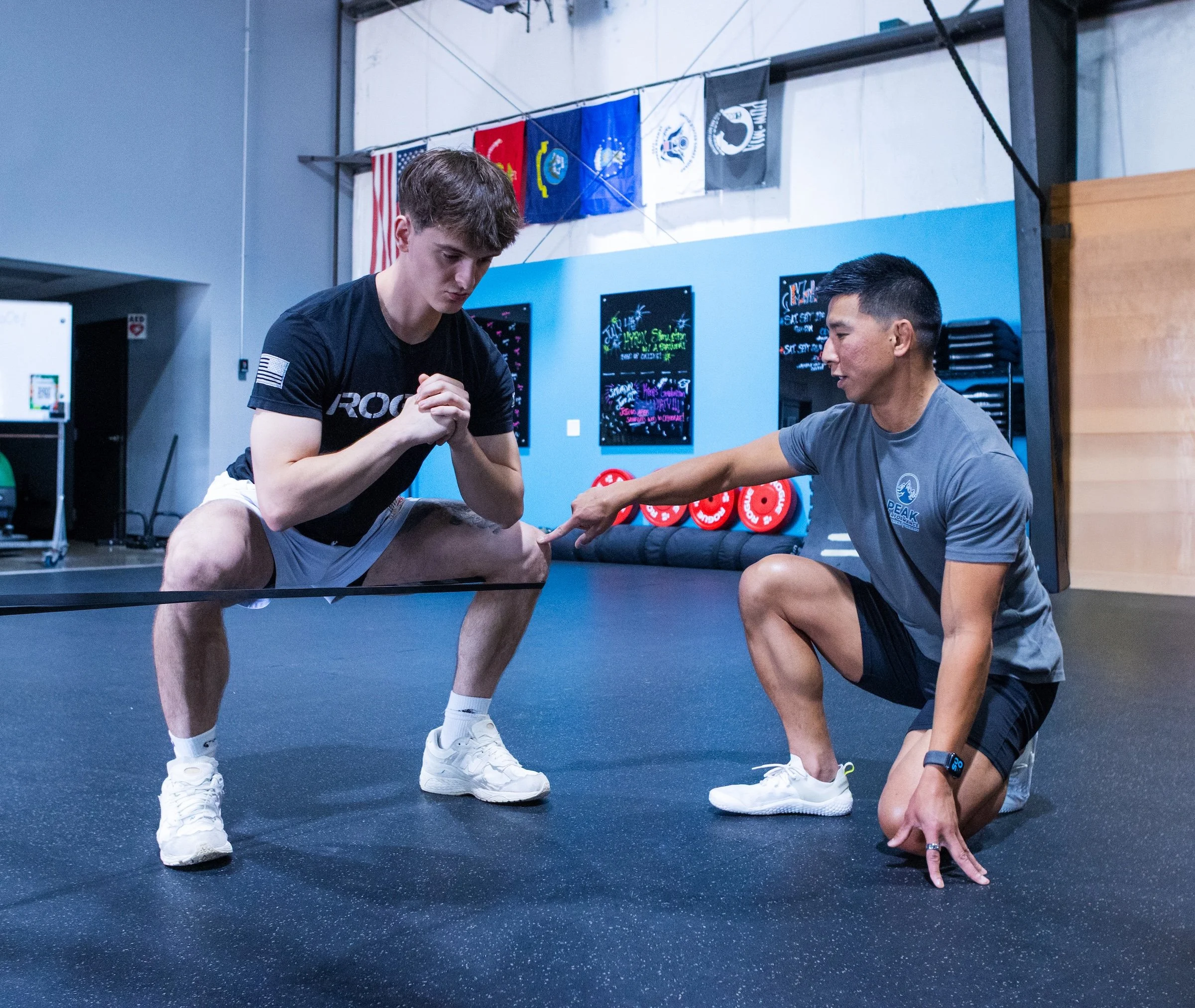 Two men practicing arm wrestling in a gym. The man on the left is young with short brown hair, wearing a black shirt and gray shorts. The man on the right has short black hair, wearing a gray shirt and black shorts. Both are focused and gripping each other's hands on a table, with flags and motivational signs in the background.