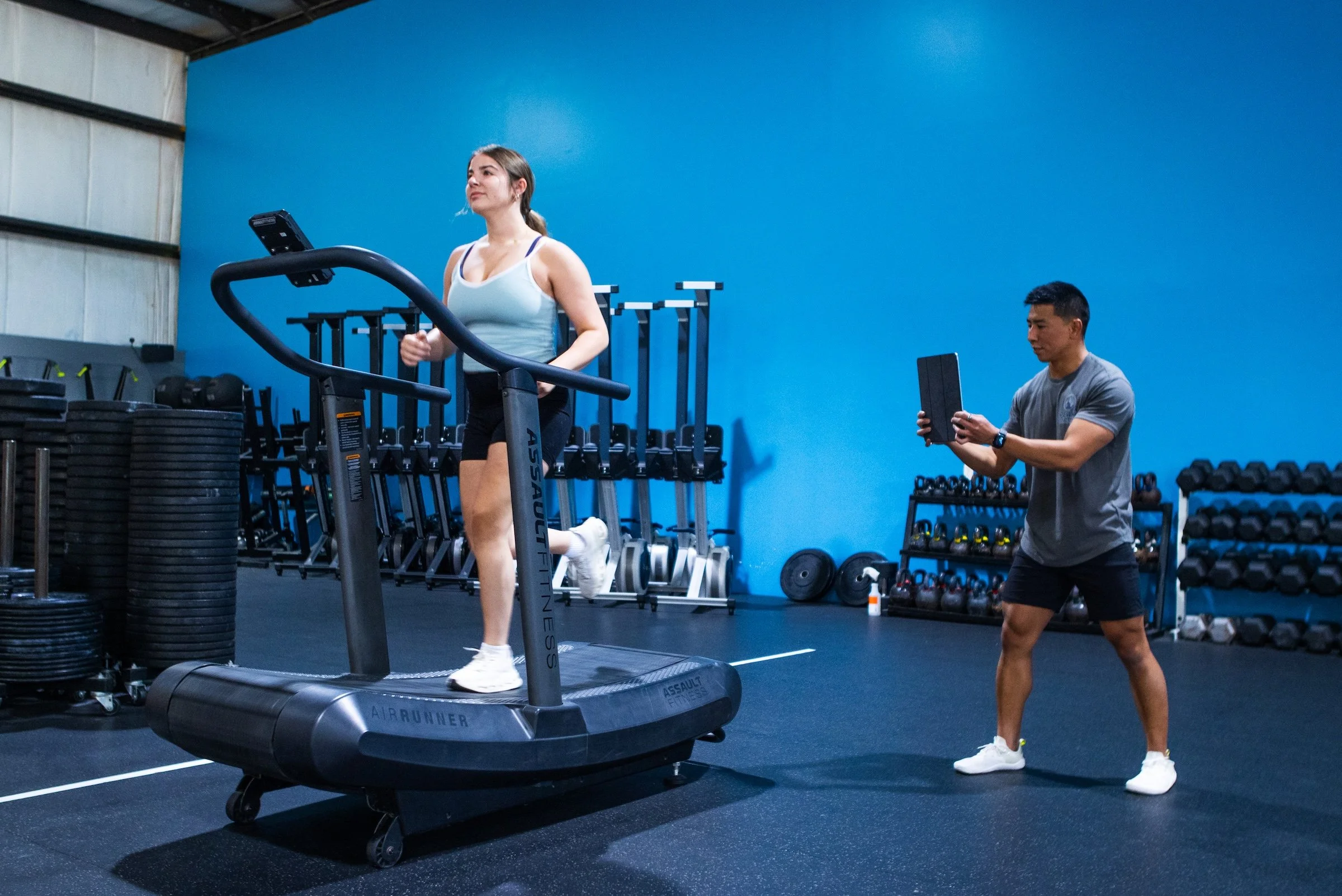 Woman running on a treadmill while a trainer supervises in a gym with blue walls, weight racks, and exercise equipment.