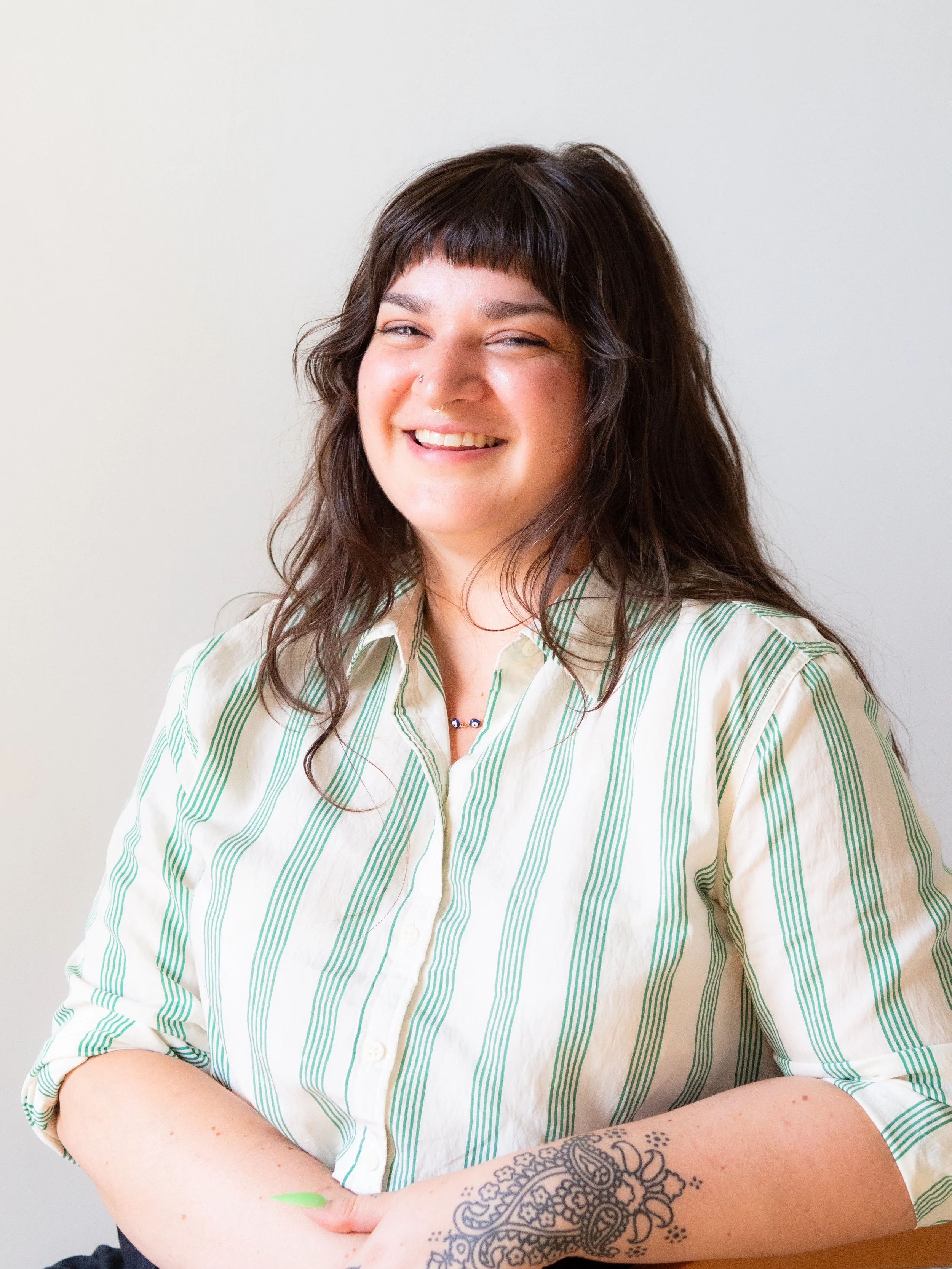 A woman with long dark wavy hair and a nose piercing smiling while wearing a beige and green striped shirt, sitting against a plain white wall.