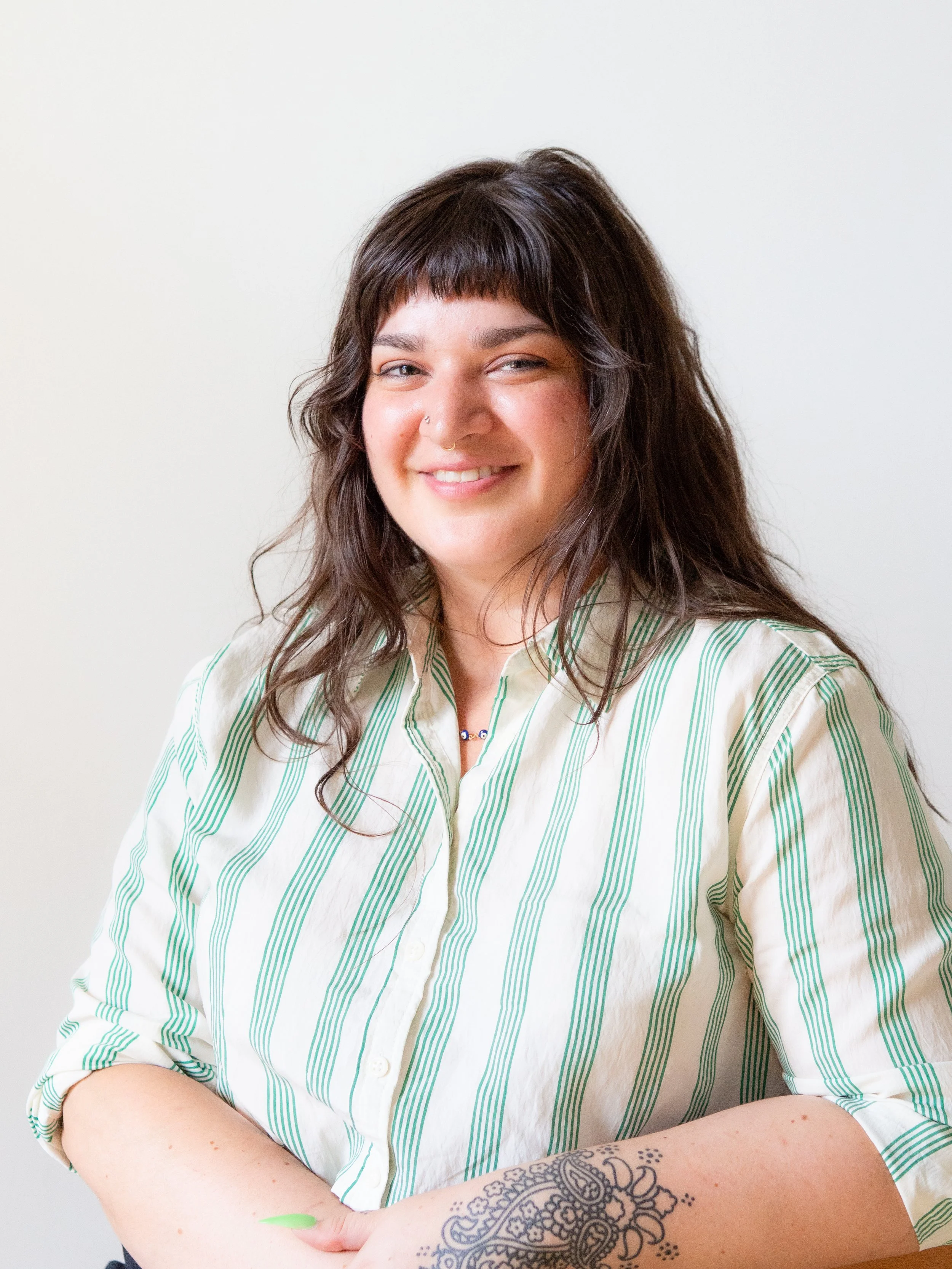 A woman with long, wavy dark hair, wearing a cream-colored shirt with green vertical stripes, smiling and looking at the camera. She has a nose ring, a septum piercing, and a tattoo on her left forearm. The background is plain white.
