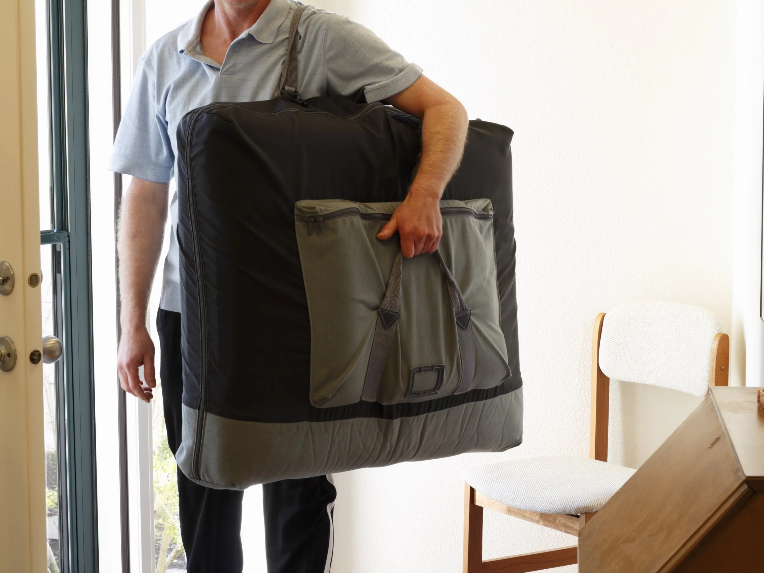A person carrying a large black and gray backpack while standing near a doorway, with a white wall and a wooden chair in the background.