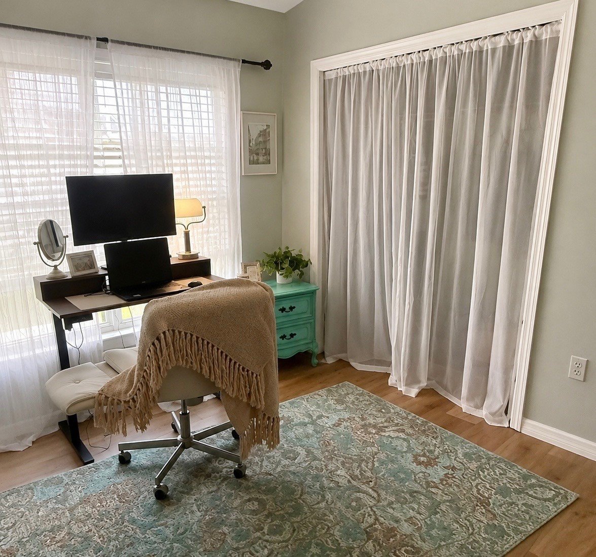 A home office with a desk holding a computer monitor, a smaller screen, a lamp, and a mirror; a beige office chair with a throw blanket; a teal side table with potted plant and photo frames; sheer white curtains over large windows and a door with gauzy curtains; a patterned area rug on hardwood flooring.