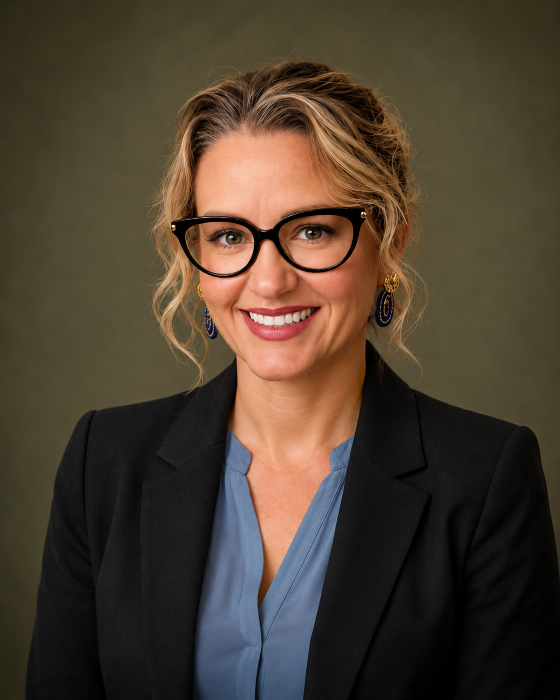 Suzana Hachey, APRN, PMHNP-BC, wearing glasses, a black blazer, and a blue blouse, smiling in front of a gray background.