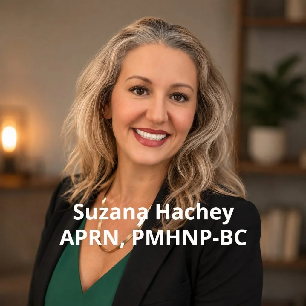 Suzana Hachey, APRN, PMHNP-BC smiling, with curly blonde hair, wearing a black blazer and green top, standing indoors with a blurred background of a bookshelf and warm lighting.