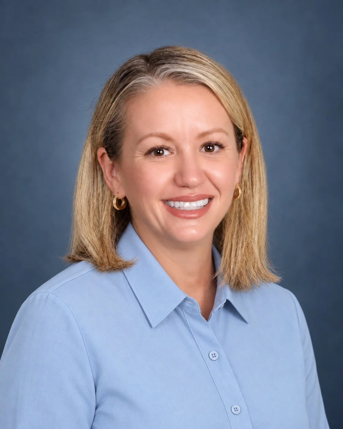 Headshot of a woman with blonde hair, wearing a light blue collared shirt, gold hoop earrings, and smiling against a solid dark background.