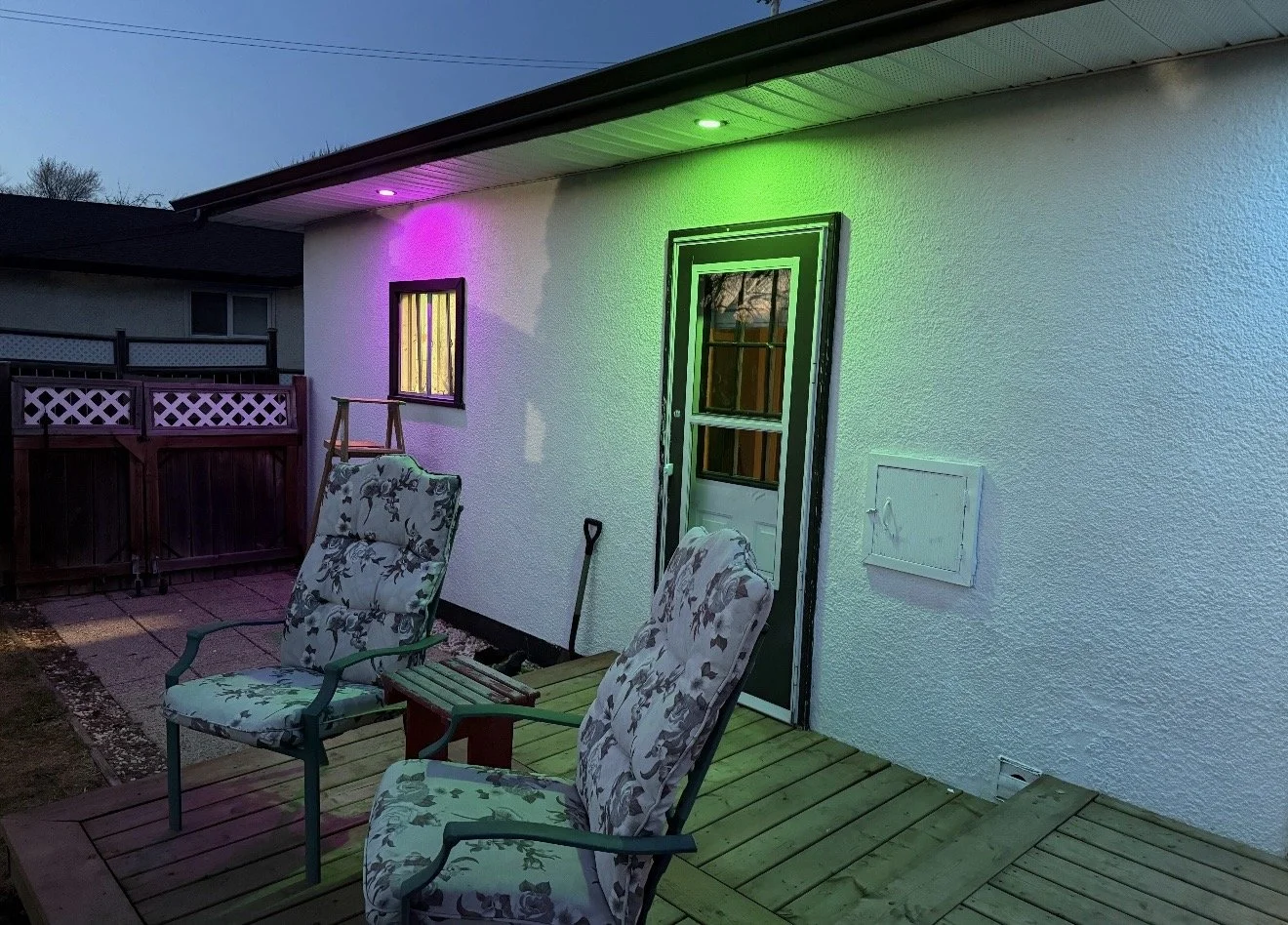 Backyard deck with two floral cushioned chairs, a small wooden table, and colorful LED lights illuminating the house exterior at dusk.