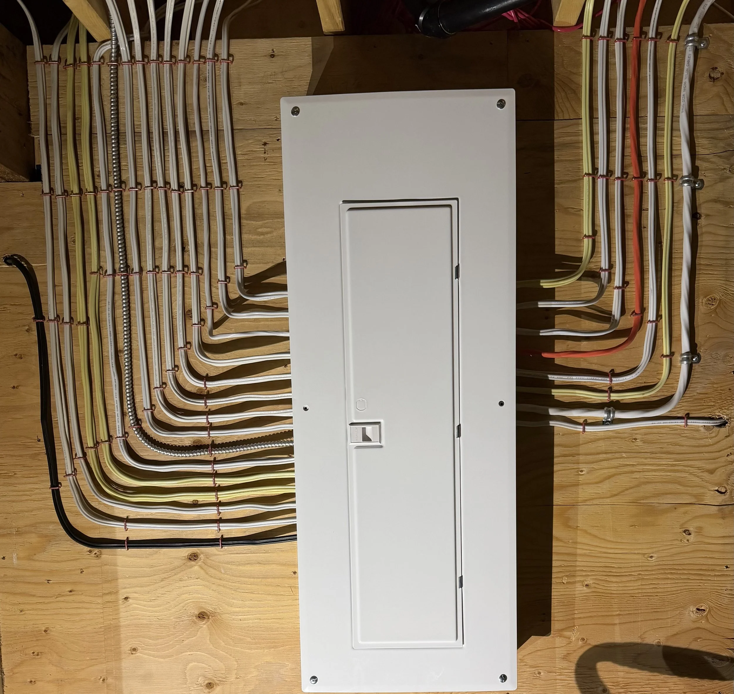 Electrical wiring and conduit organized on a wooden wall, with a white electrical panel and metal connectors.