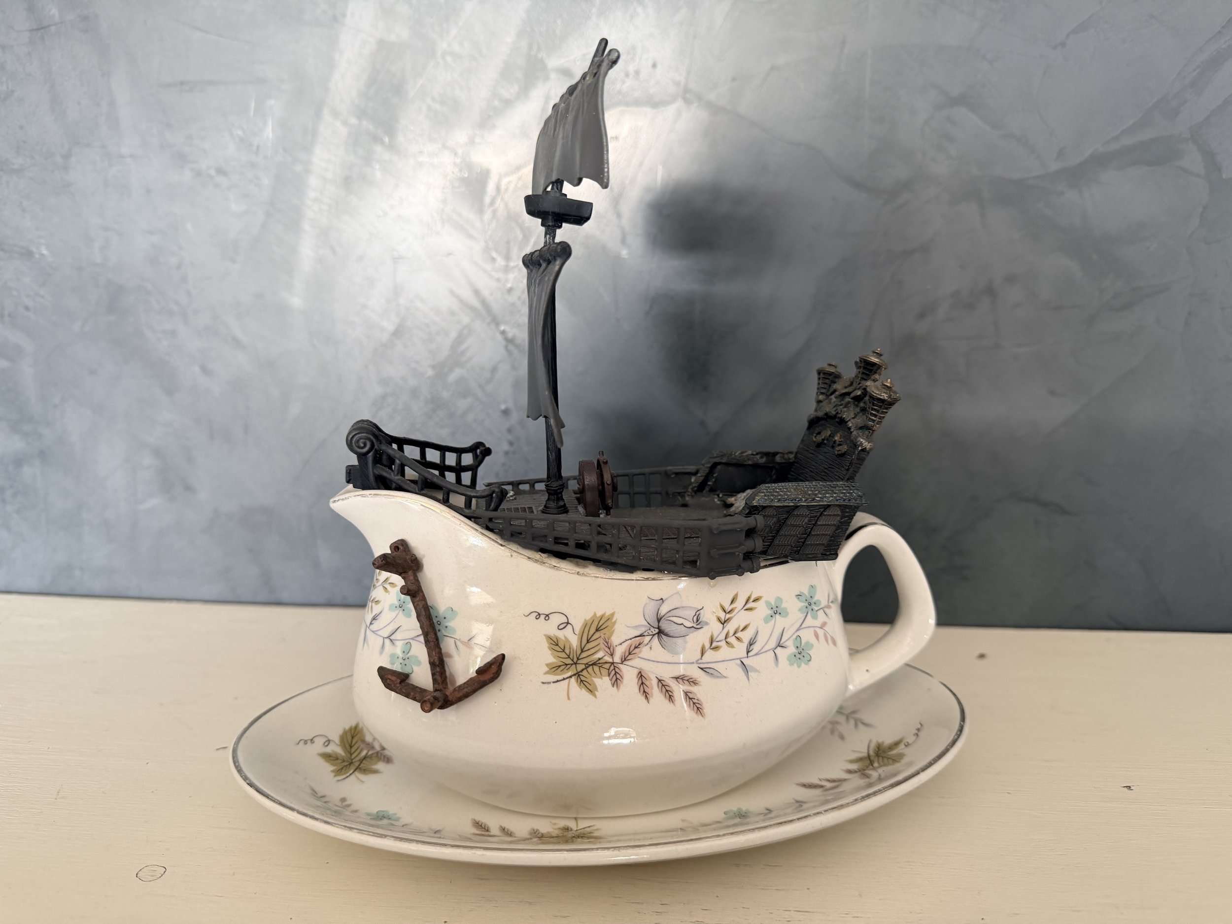 A teapot with a floral pattern on a white surface, containing a miniature wrought iron shopping cart, an anchor, and some decorative items.