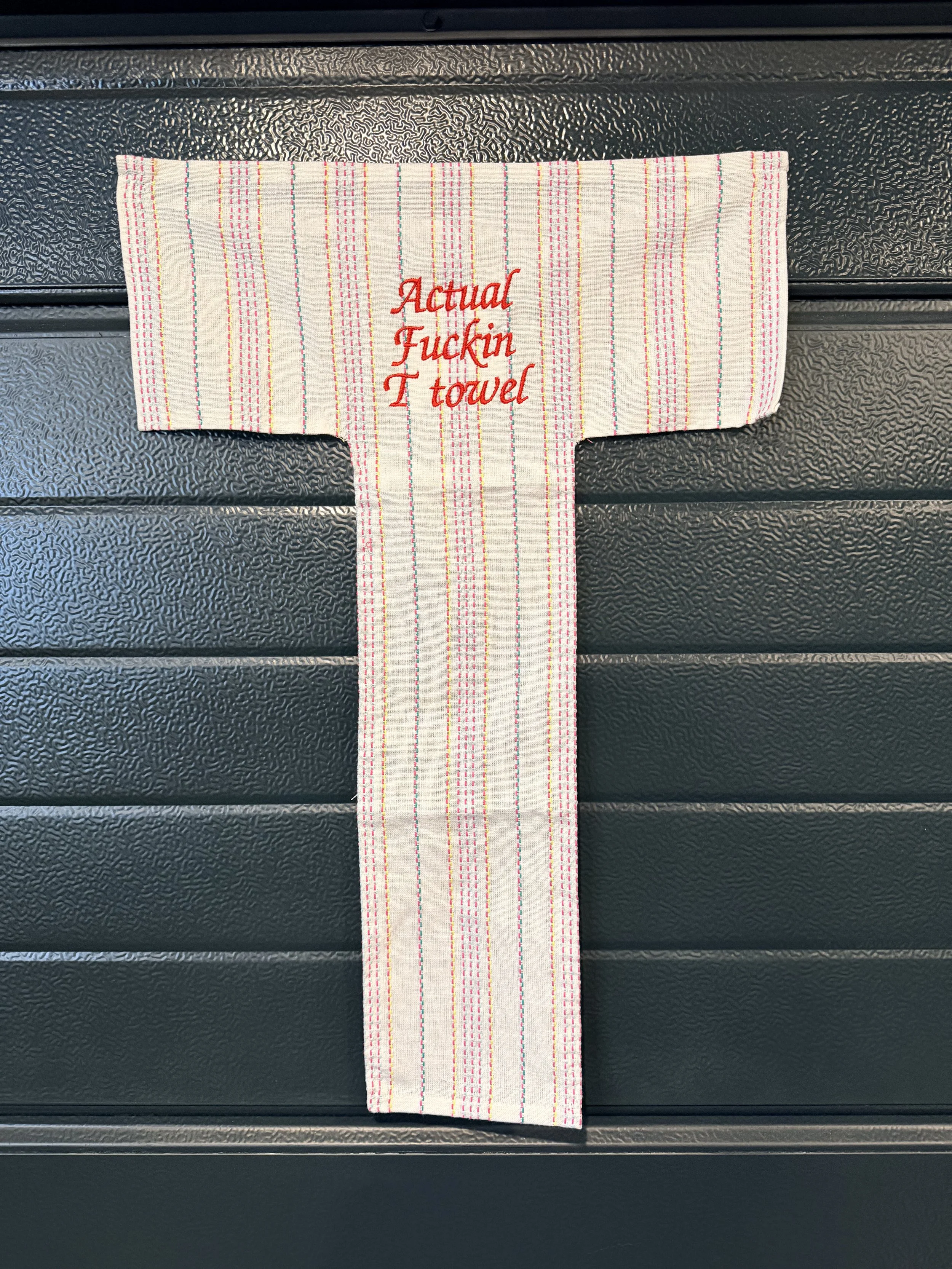 A white dish towel with multicolored vertical stripes and red embroidery that reads 'Actual Fuckin Towel', placed on a black textured surface.