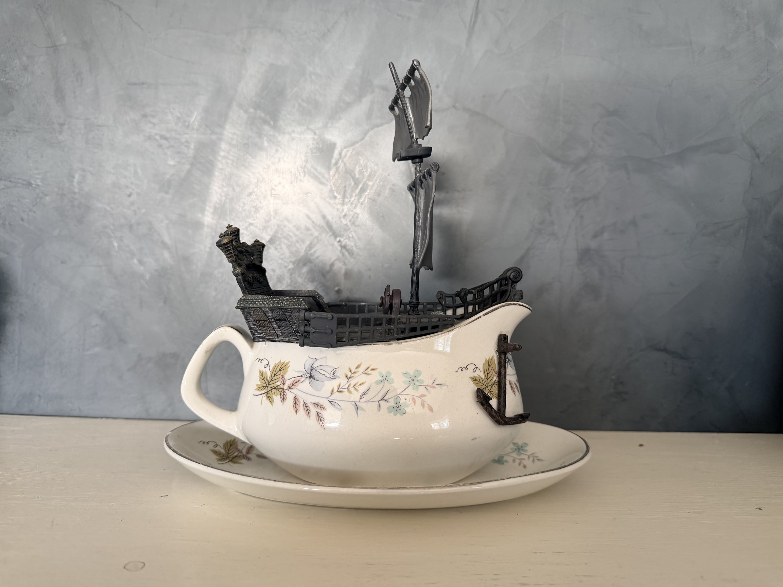 A vintage teapot white with floral patterns, topped with a miniature shopping cart, a small piece of metal pipe, and a black sculpture of a hand, on a table with a gray textured background.