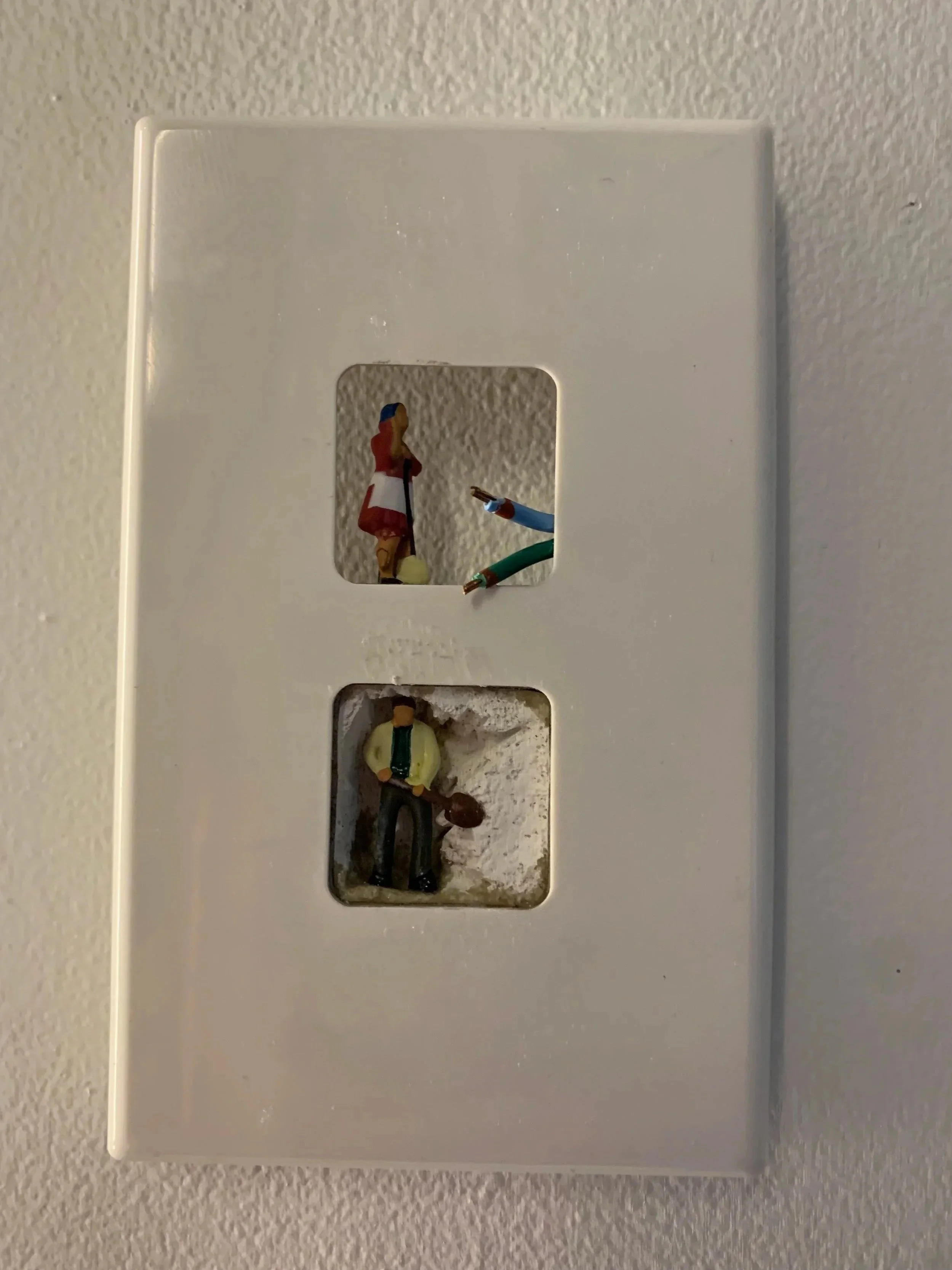 Decorative light switch cover plate with miniature figures of three people, one woman and two men, inside the openings.