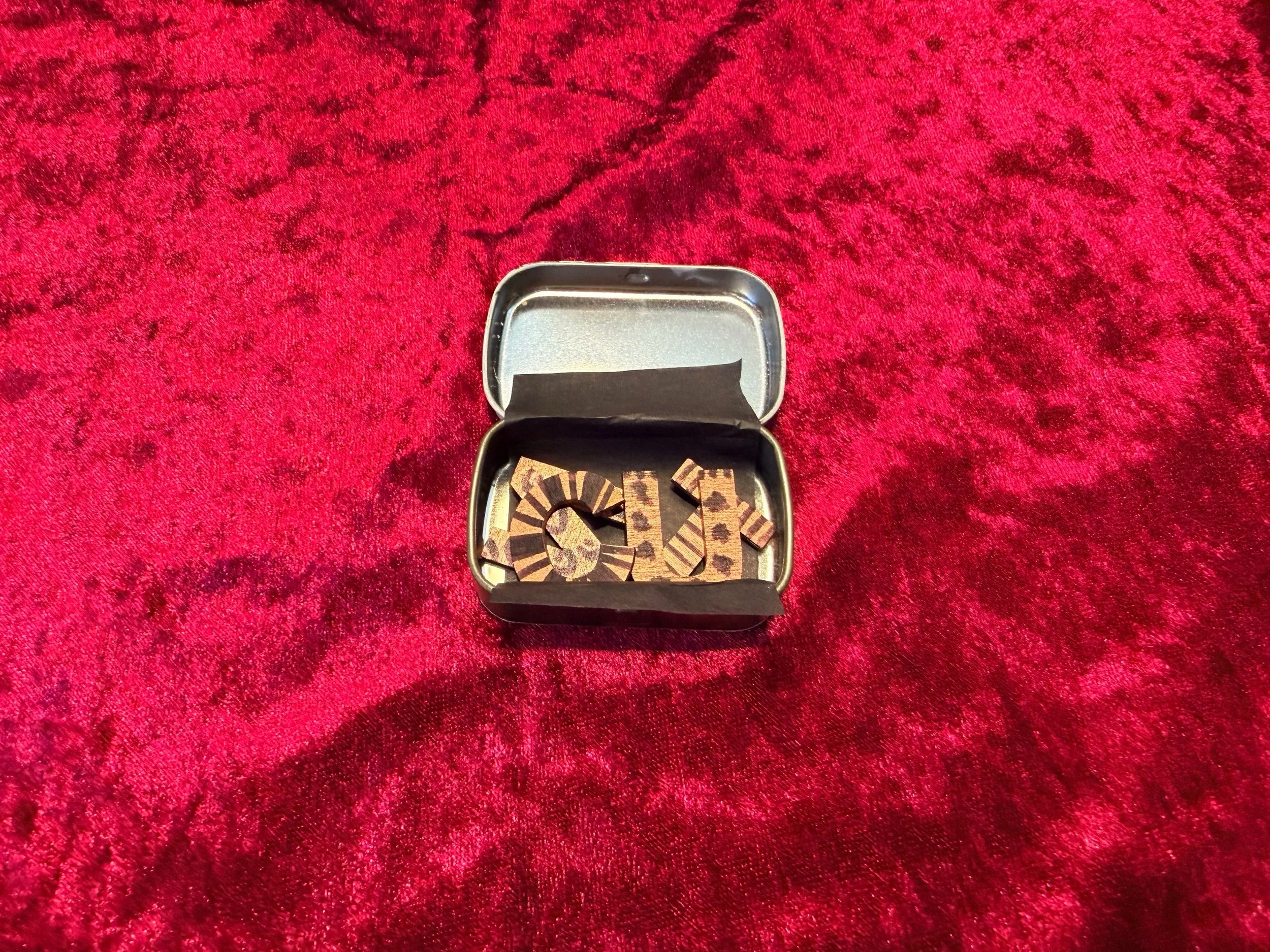 A small metallic container holding several small wooden or cardboard pieces with various black patterns and cuts, placed on a red velvet textured surface.
