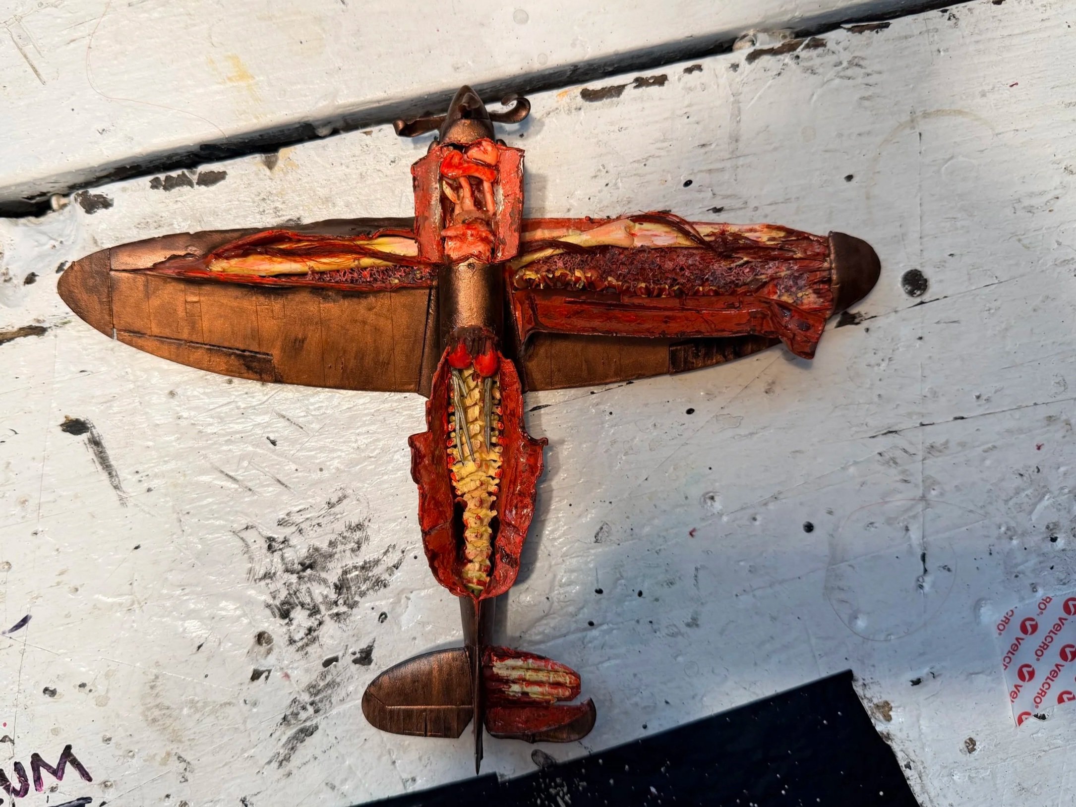 A metal sculpture resembling a dragonfly laid on a white, slightly distressed surface with black marks, scratches, and ink stains.
