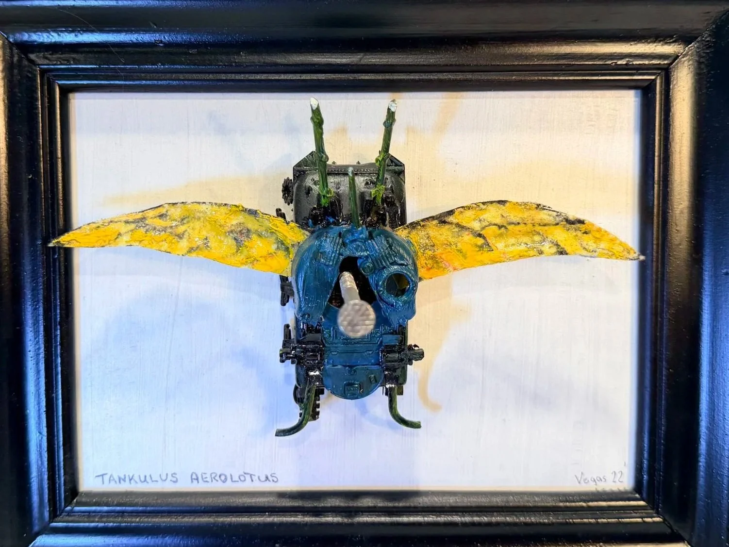 A framed artwork of a mechanical insect with yellow and black wings, a blue head, and green antennae, on a white background. The insect has a cylindrical body and detailed metallic parts. The artist's signature and date are visible at the bottom.