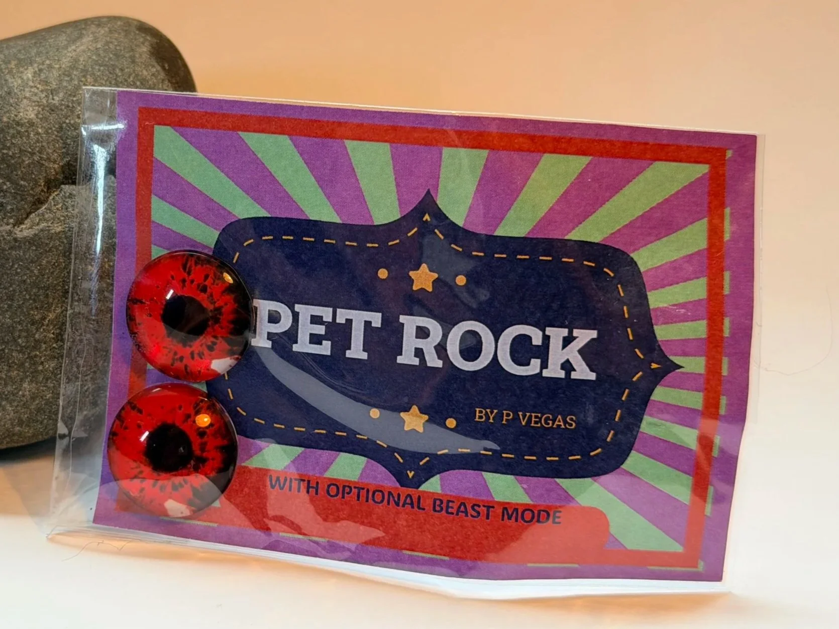 Package of toy eyes labeled 'Pet Rock' with three red and black plastic eyes, inside a clear plastic bag. The background features a purple, green, and red design with the words 'Pet Rock' and mentions optional beast mode.