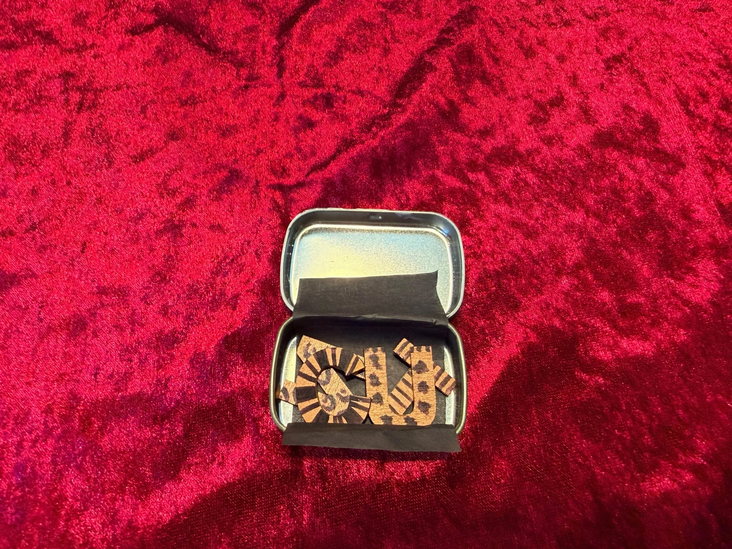 A small rectangular metal tin with an open lid, containing chess pieces, sits on a red textured fabric background.