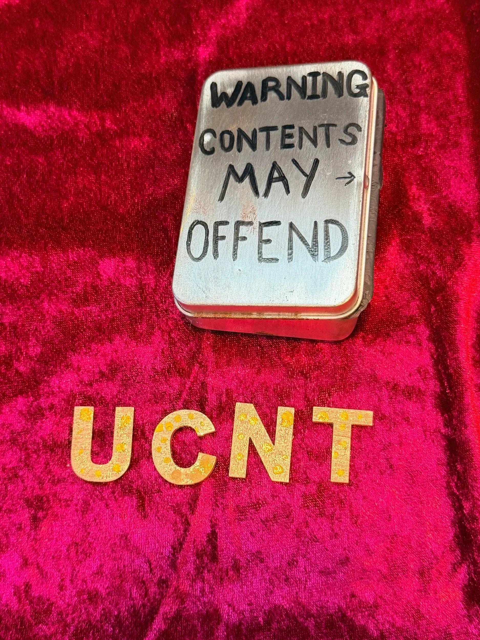 A metallic square container with a warning label in black letters indicating contents may offend, placed on a pink, textured surface with partially visible decorative letters spelling 'JUCIENT' underneath.