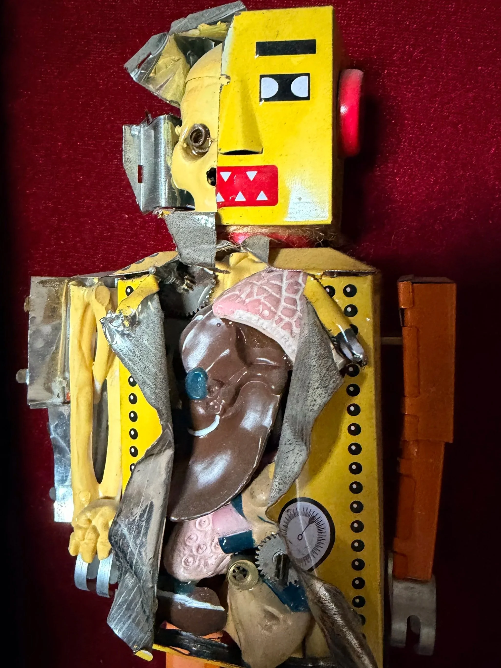 A broken toy robot with a yellow face, red and black eyes, and a large open mouth with white teeth, lying on a red surface surrounded by torn metal and plastic pieces.