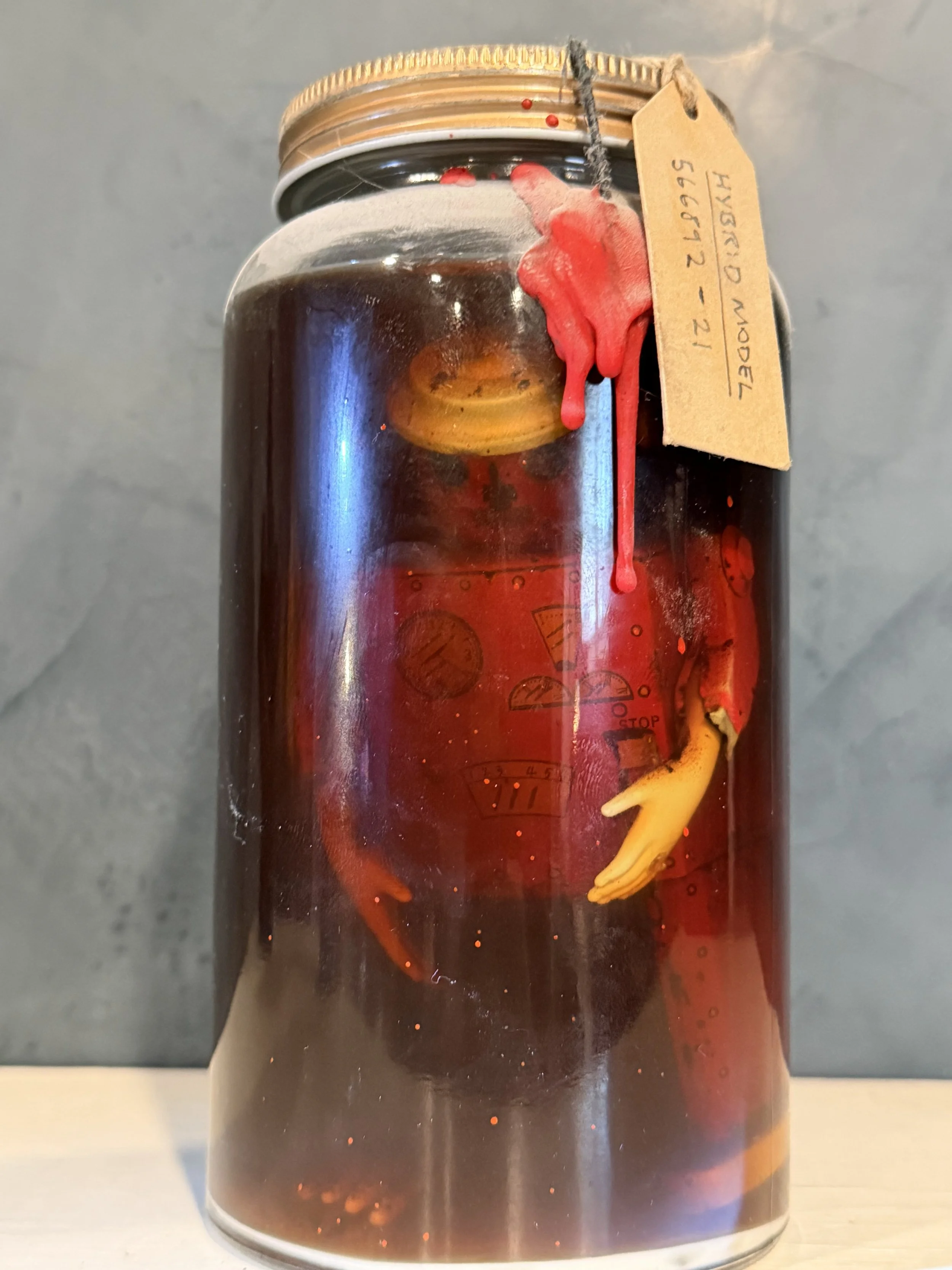 A jar with a metal lid containing a dark liquid, with a yellow rubber glove attached to the side, and a paper tag tied to the lid.