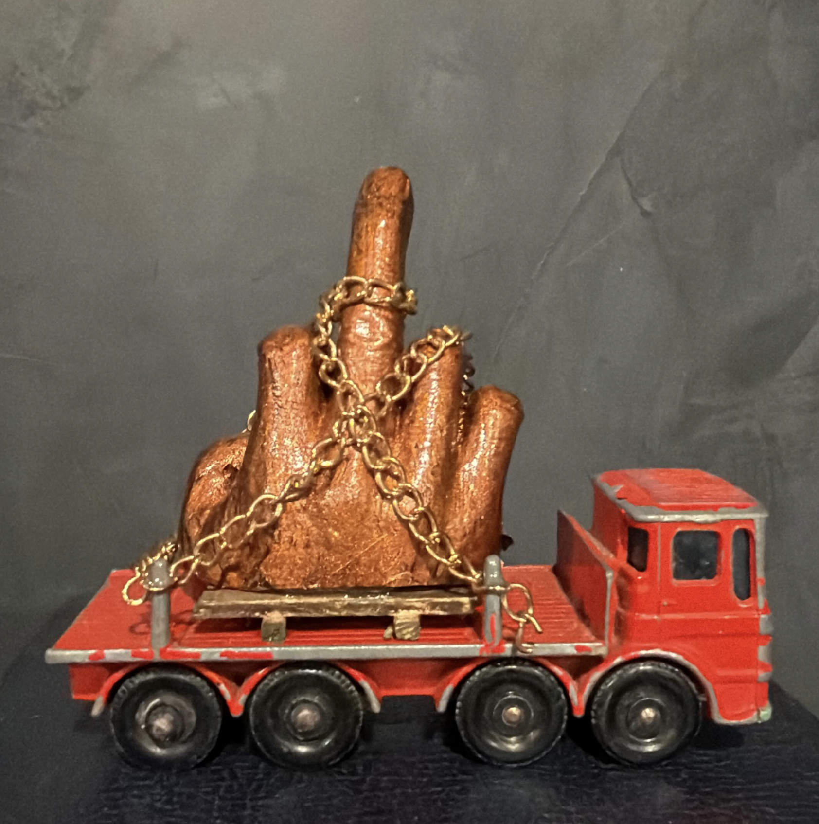 A model train with a red locomotive carrying a large, golden-brown statue of a hand with two fingers raised, adorned with a gold chain, against a dark background.