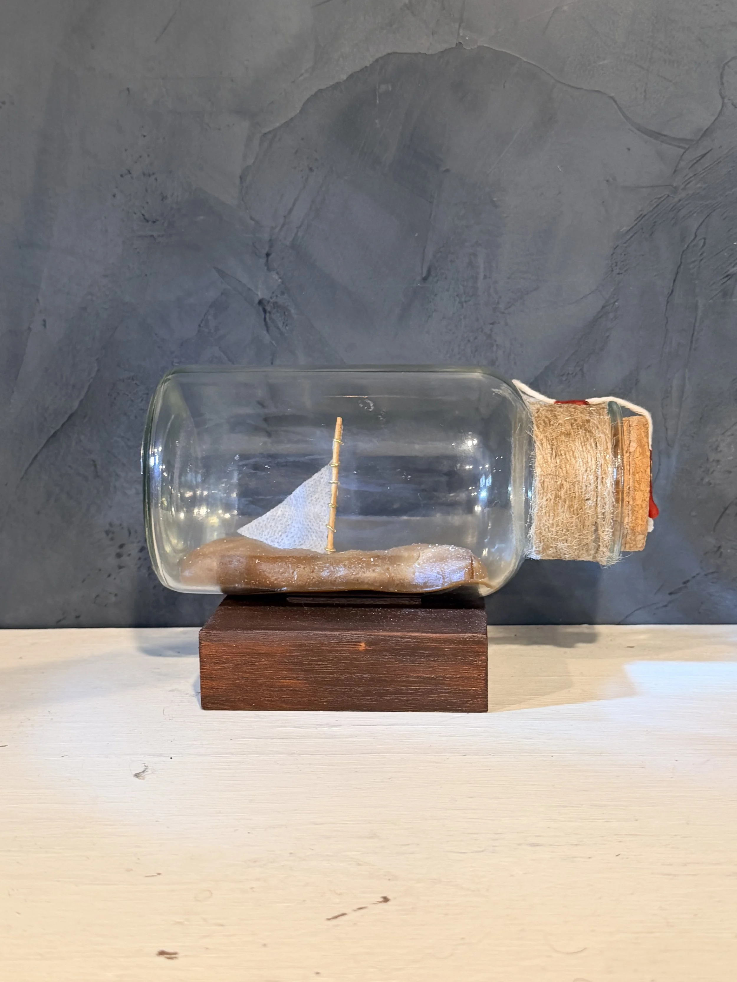 A glass container with water, a piece of paper towel, and a wooden stick inside, placed on a wooden base, on a surface with a dark textured wall background.