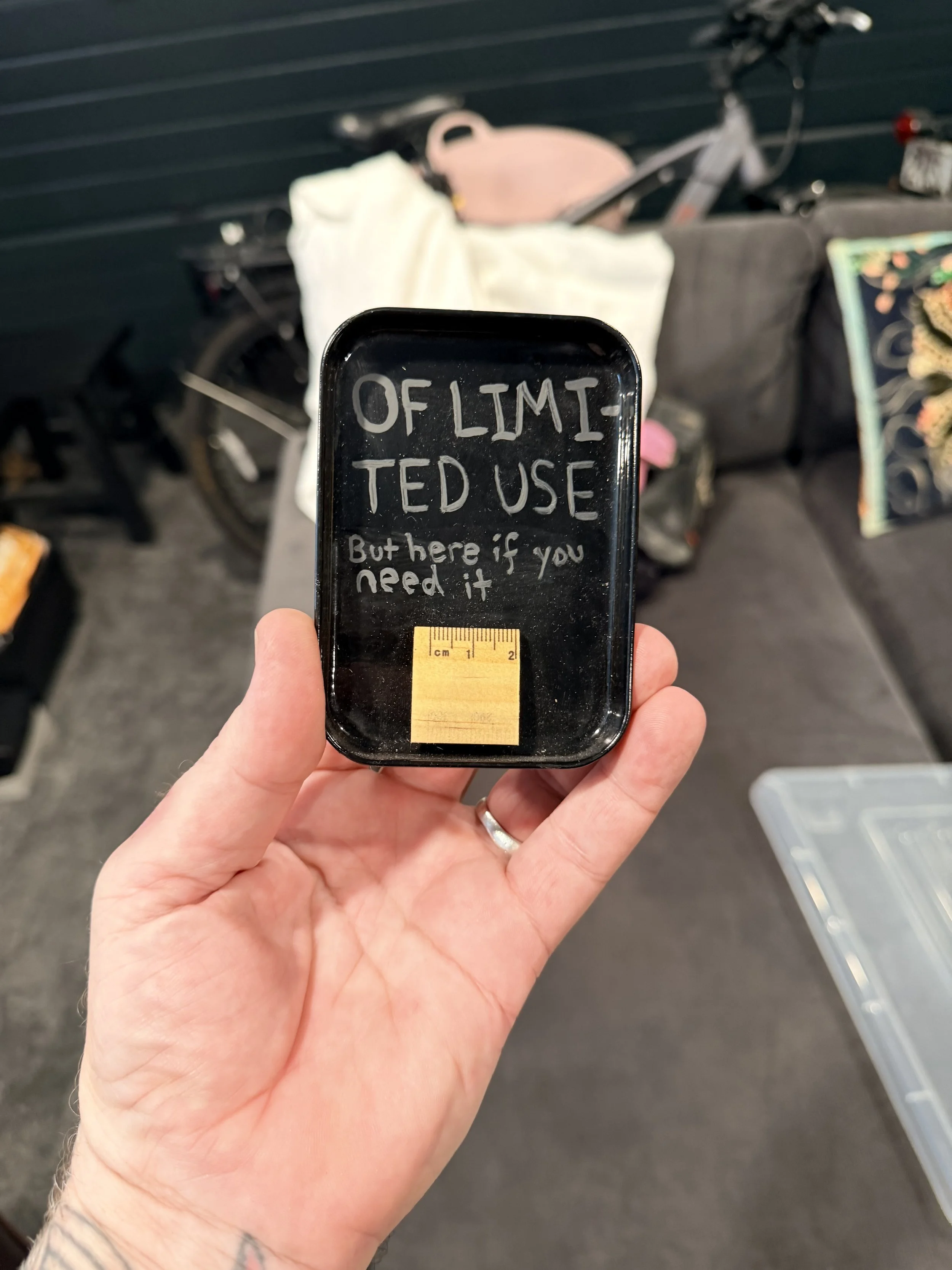 A hand holding a small black tray with white handwritten text that reads 'OF LIMITED TED USE But here if you need it'. In the background, there is a bicycle with a basket containing various items, a black bag, and a gray wall.