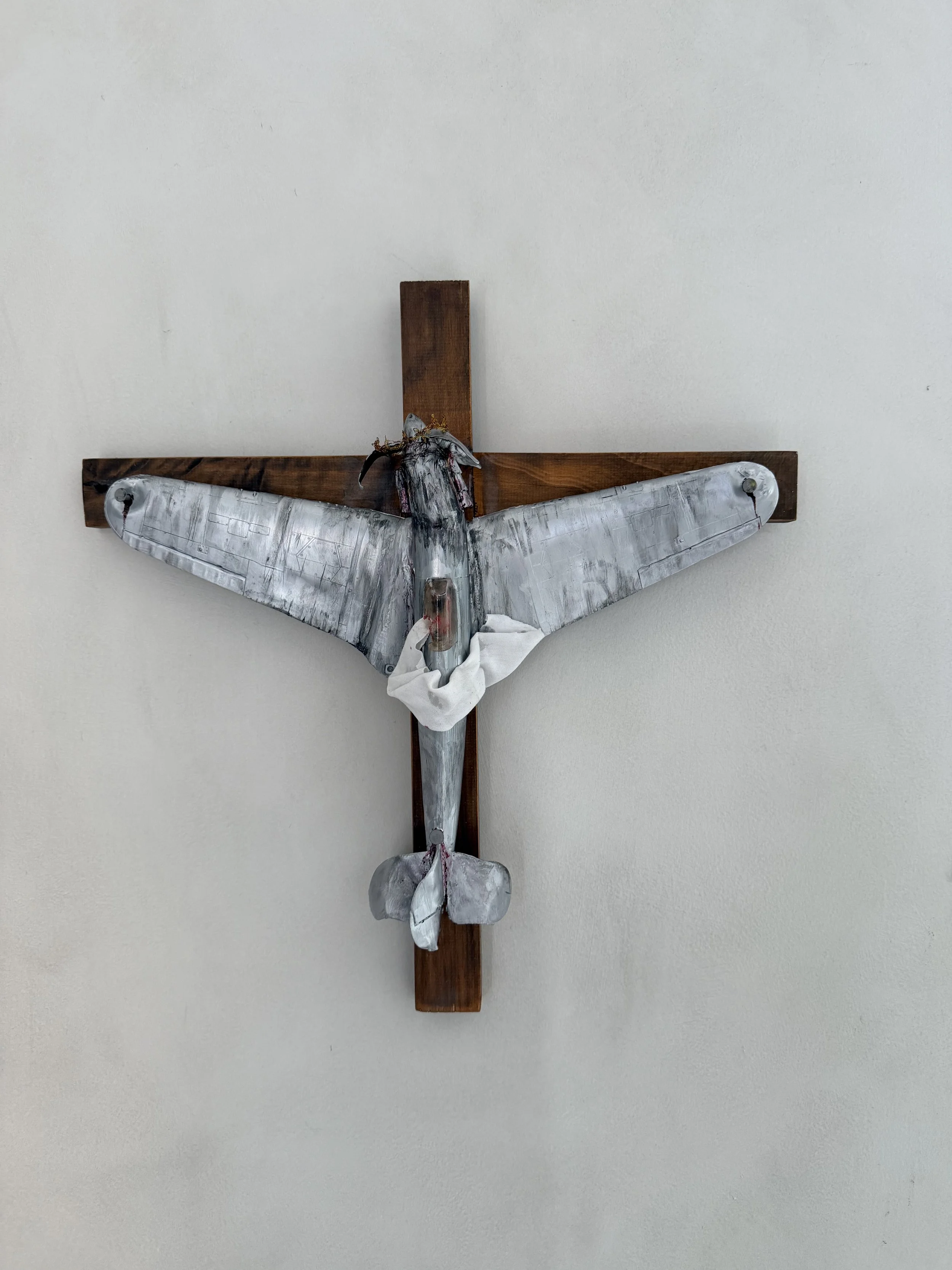 A metal moth sculpture mounted on a wooden cross on a plain white wall.