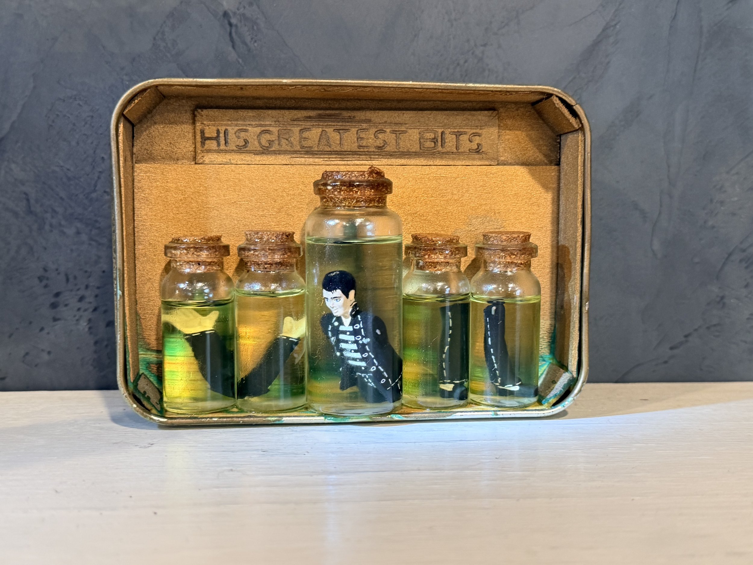 A cardboard box with the phrase 'HIS GREATEST BITS' engraved inside contains six small glass bottles with cork stoppers and a small animated figure of a man in a striped shirt and black leather jacket.