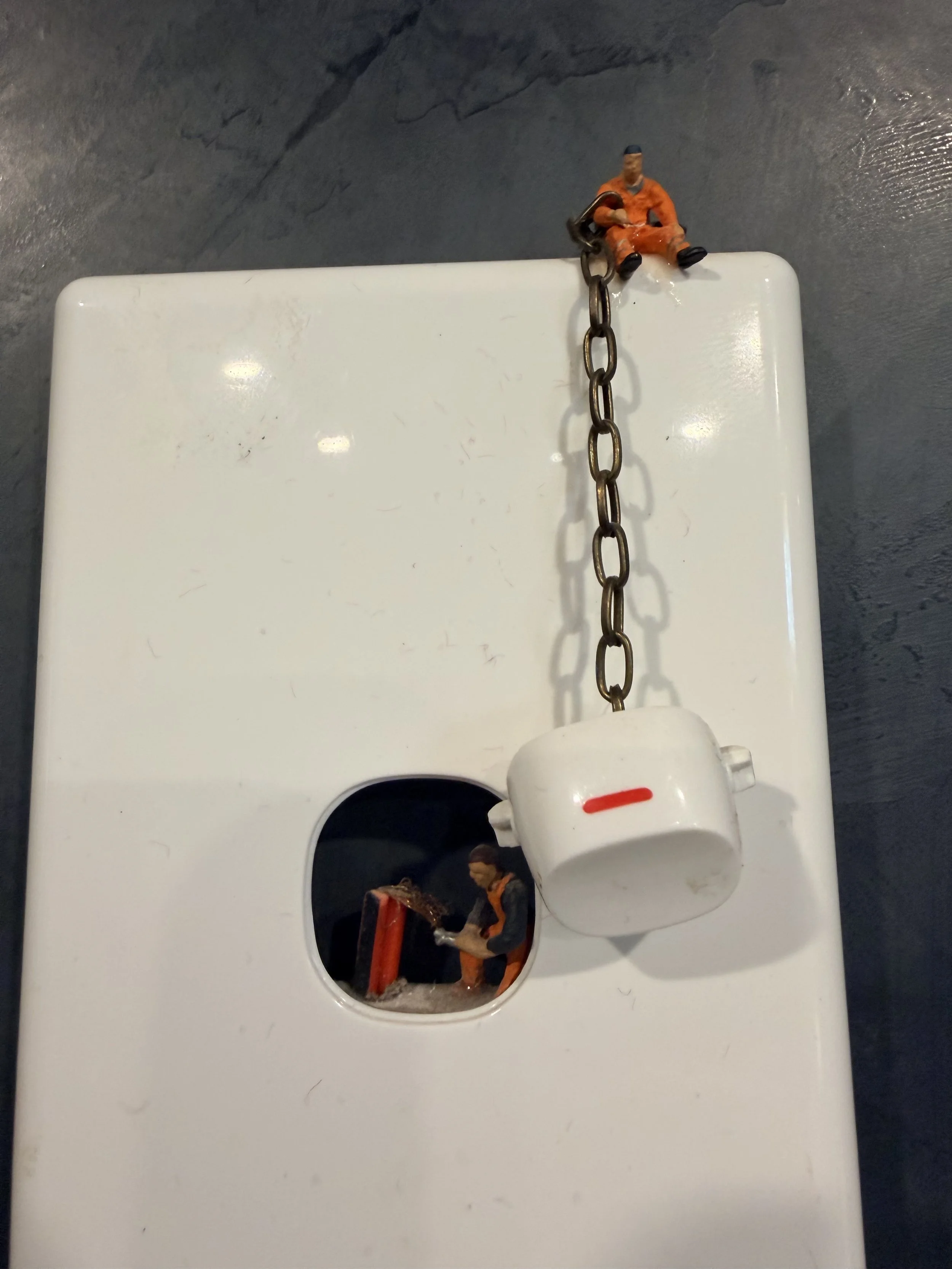 A white electrical switch cover with two toy figures, one holding a red object, and a chain with a small orange figure sitting on top of the chain. The background is a gray wall with a textured surface.